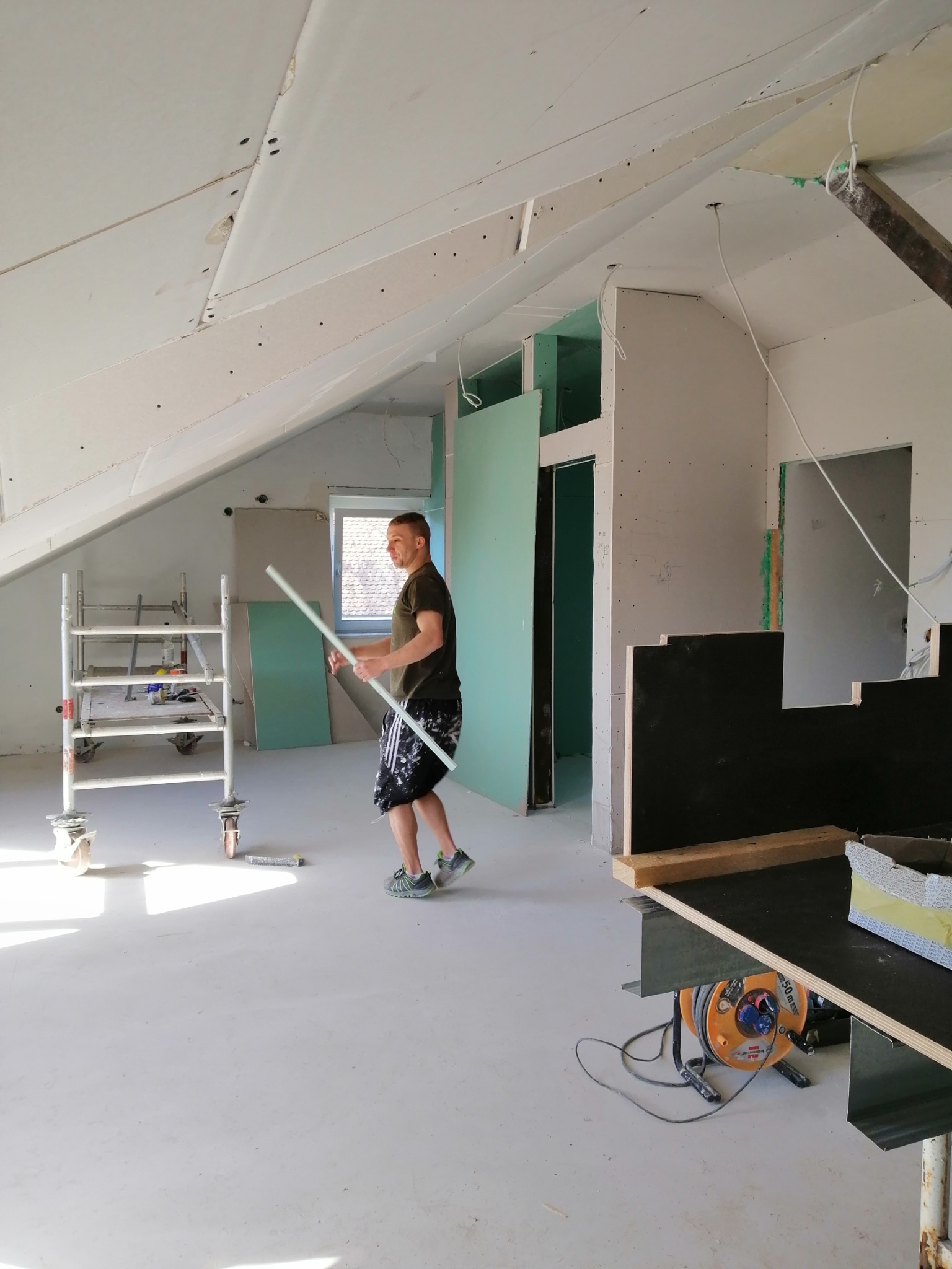 Prace wykończeniowe poddasza w Groß-Rohrheim: pracownik z profilem aluminiowym na tle rusztowania i ścian z płyt gipsowo-kartonowych przed szpachlowaniem.