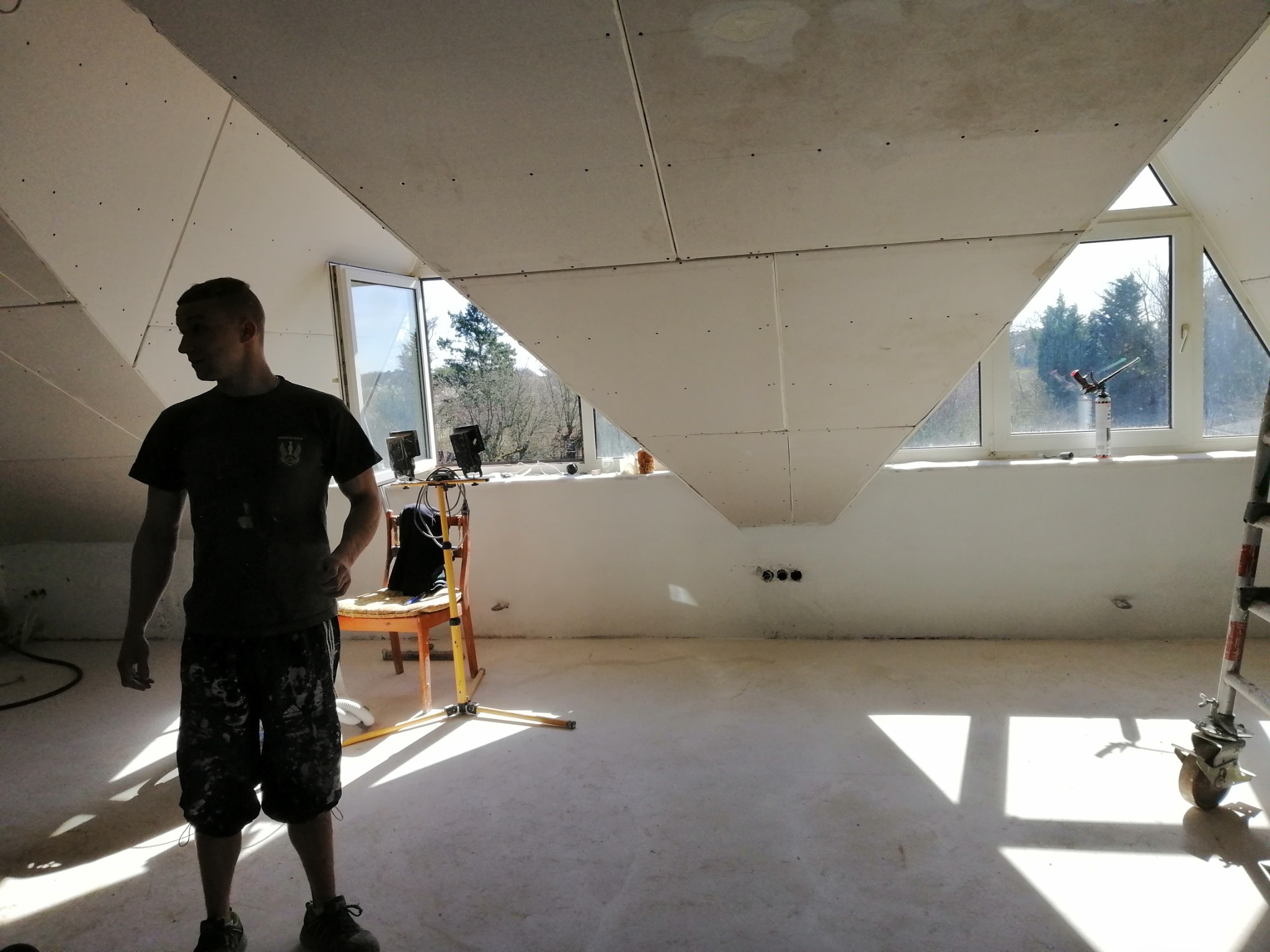 Montaż płyt gipsowo-kartonowych na poddaszu, widoczny pracownik w odzieży roboczej, okna dachowe i częściowo zamontowane płyty, widoczne narzędzia i elementy konstrukcyjne.