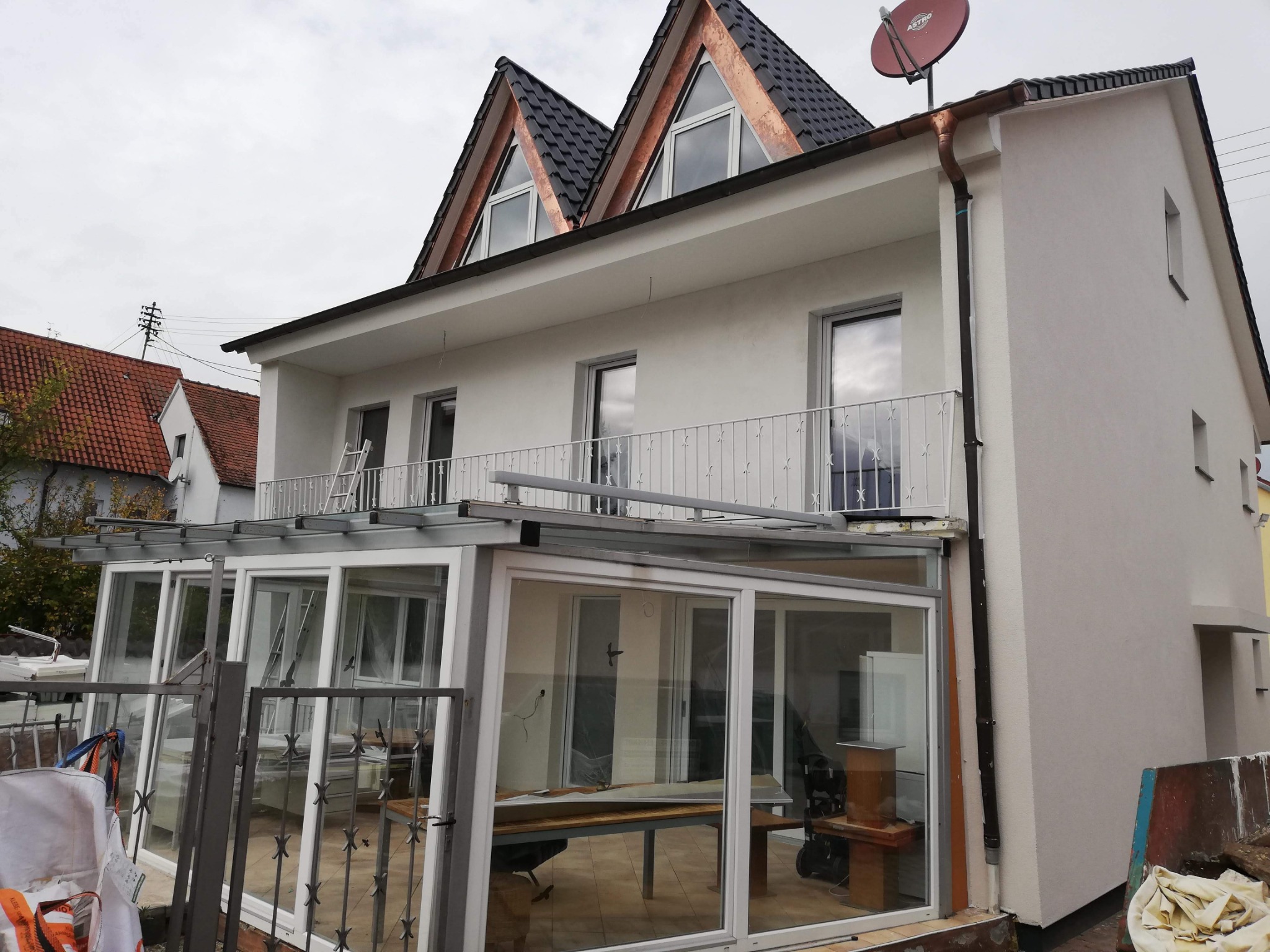 Dwupiętrowy dom z przeszkloną oranżerią, świeżo otynkowaną elewacją i miedzianymi elementami dachu, widziany z zewnątrz, z widoczną drabiną na balkonie i elementami remontowymi w oranżerii.