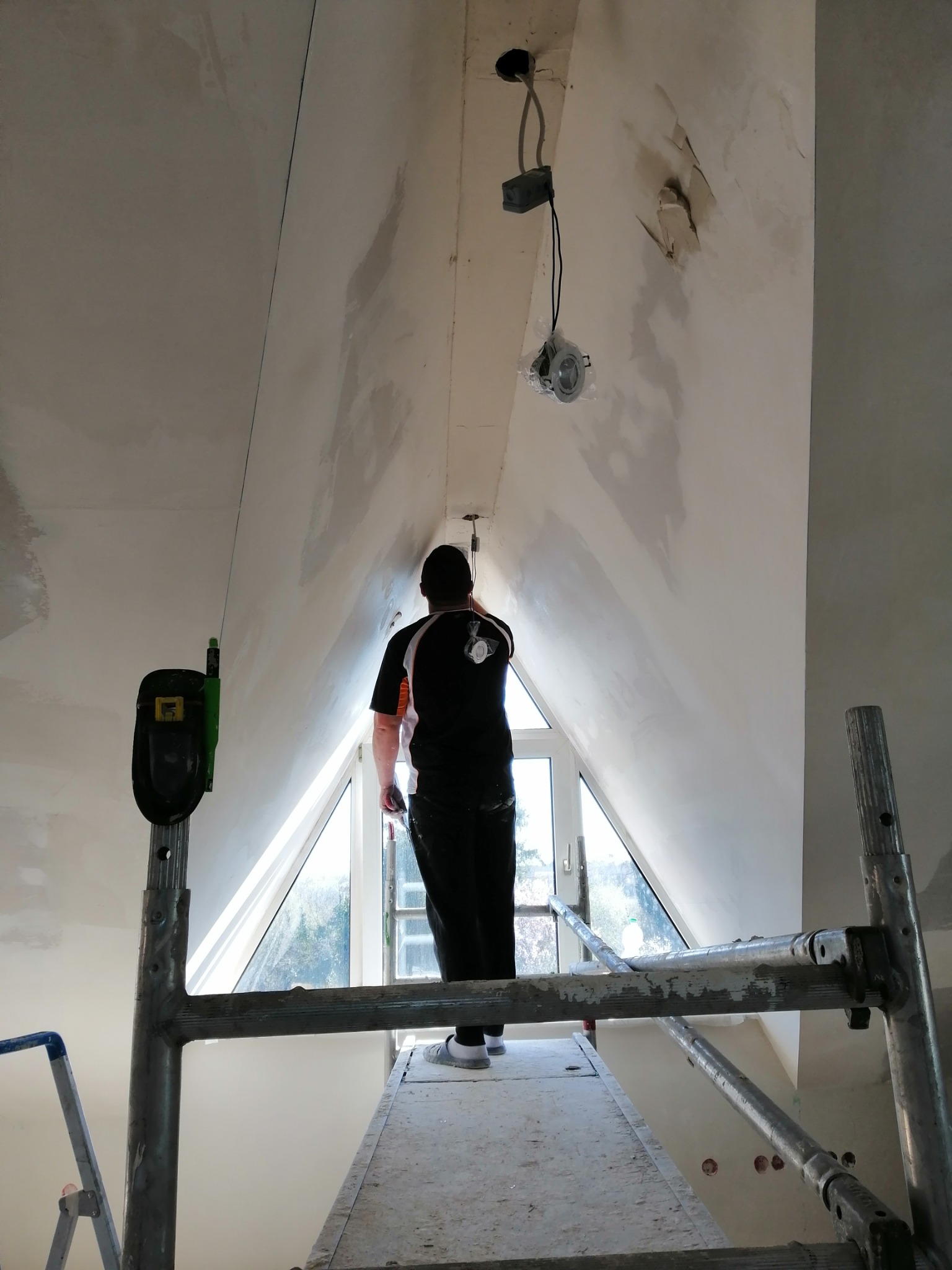 Pracownik na rusztowaniu, przygotowujący ścianę poddasza do malowania, widoczne ślady po szpachlowaniu, okno trójkątne w tle, przewody elektryczne wystające z sufitu.