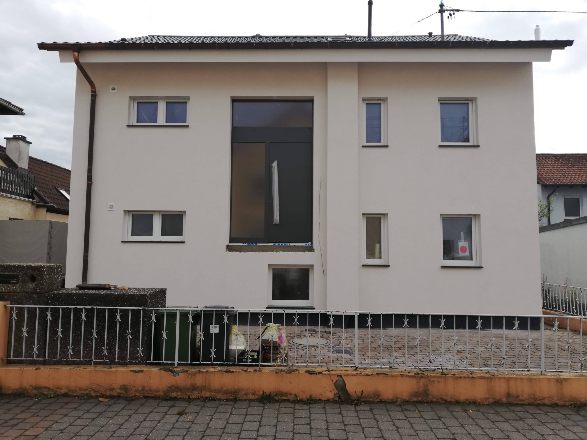 Odnowiona elewacja dwukondygnacyjnego domu z nowymi oknami, z częściowo zdjętą folią ochronną z jednego okna, widoczne ślady prac wykończeniowych i śmietniki przed domem.