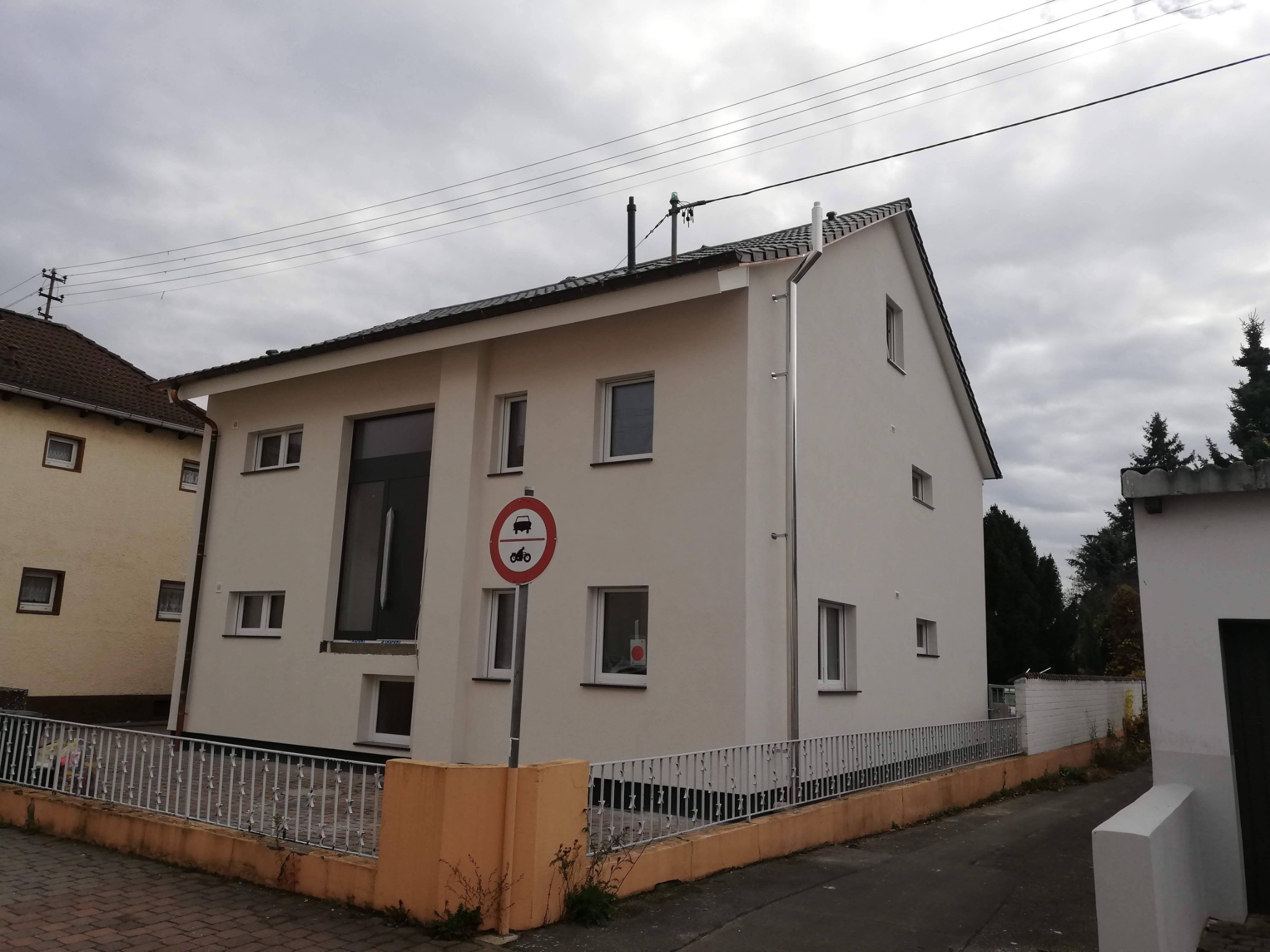 Świeżo ocieplony, dwukondygnacyjny dom z białą elewacją, widoczny znak zakazu wjazdu dla samochodów i motocykli, metalowe rynny i ciemna dachówka.