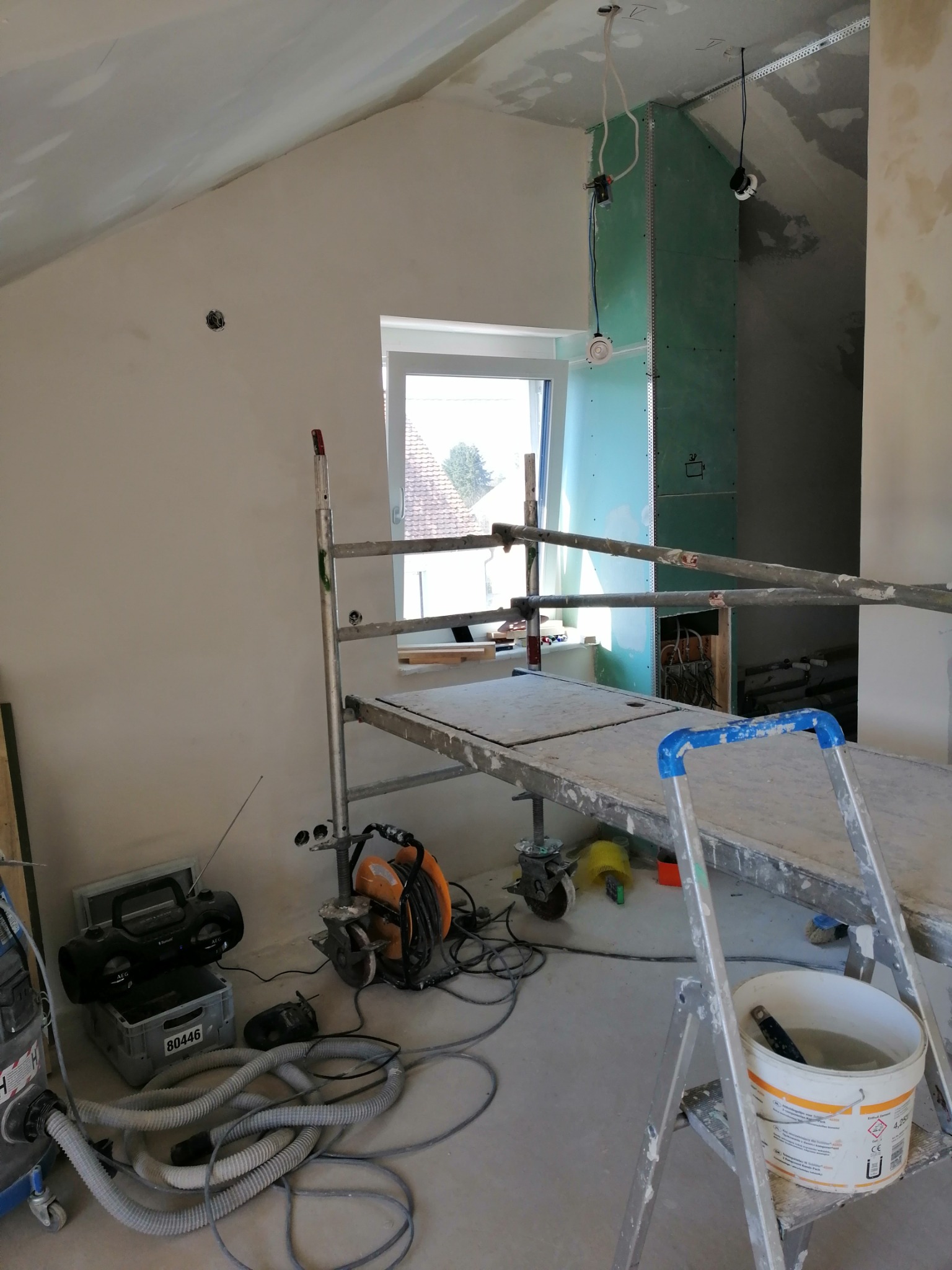 Wnętrze remontowanego poddasza z oknem, widoczne rusztowanie, drabina z wiadrem farby, agregat malarski i płyty gipsowo-kartonowe na ścianie.