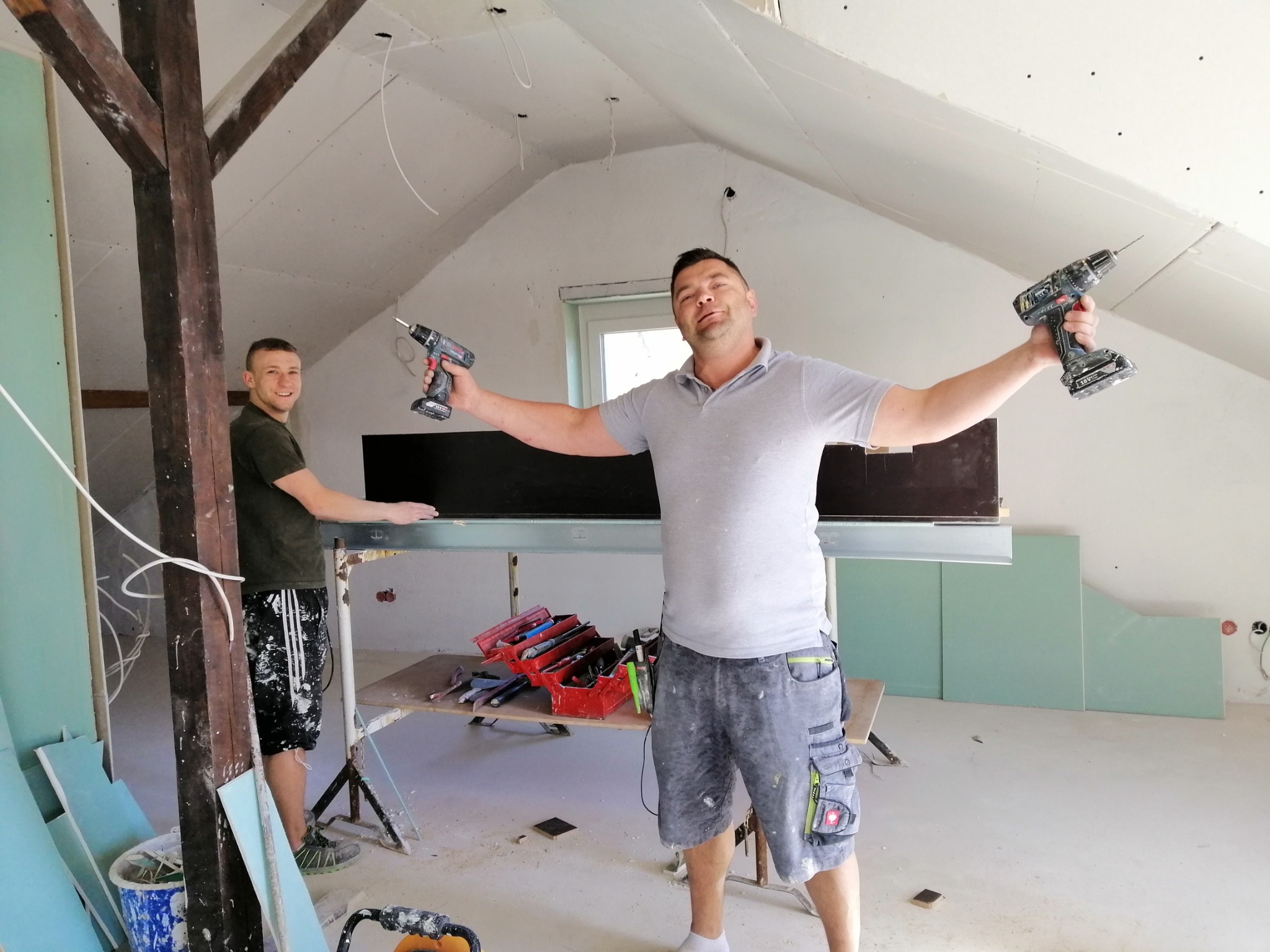 Dwóch mężczyzn w trakcie montażu płyt gipsowo-kartonowych na poddaszu, jeden z wiertarkami w rękach, drugi przy stelażu, widoczne płyty, narzędzia i drewniany słup.
