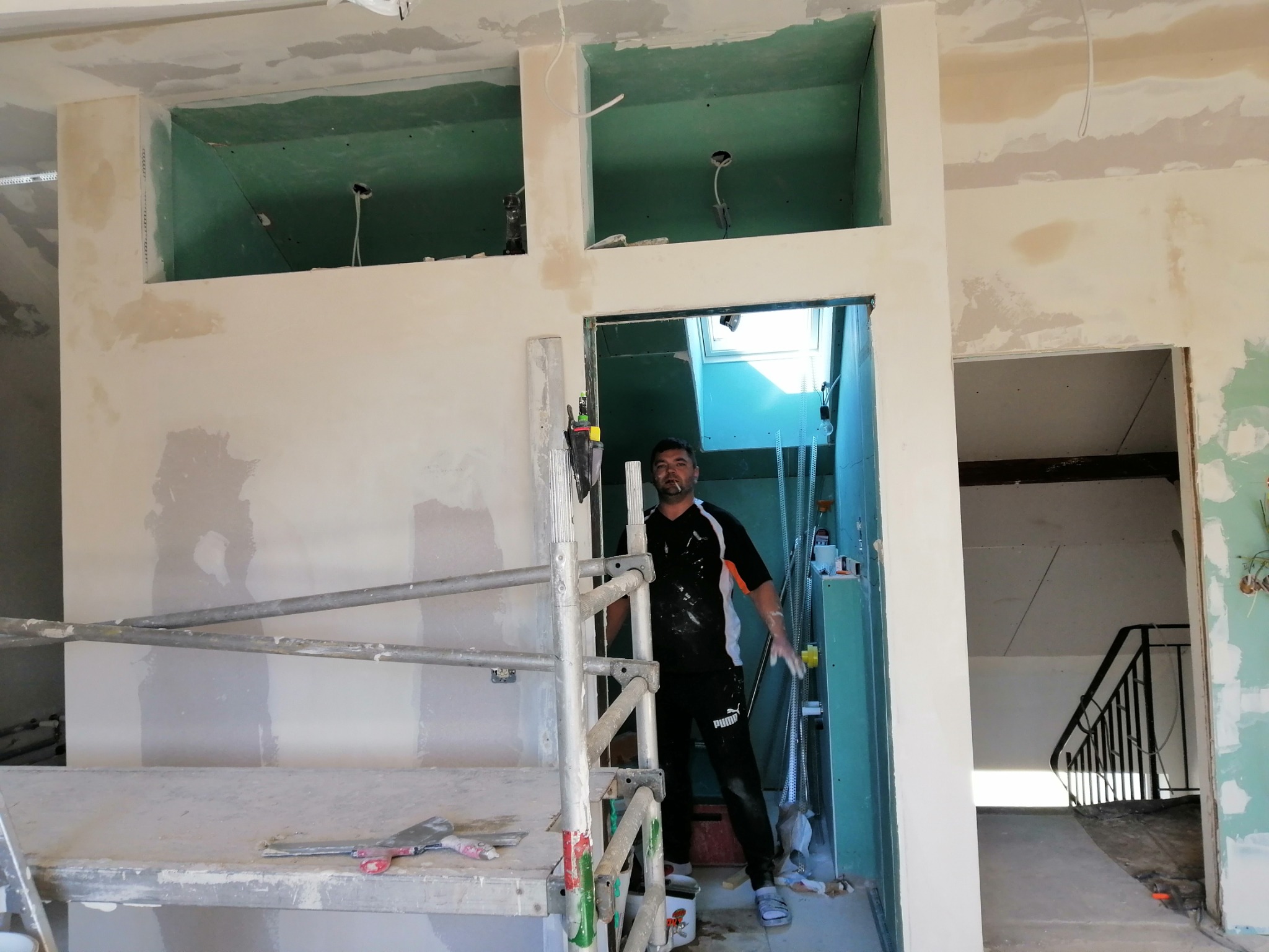 Pracownik na rusztowaniu podczas szpachlowania ścian w pomieszczeniu z przygotowanymi otworami w ścianach, widoczne narzędzia i schody w tle.