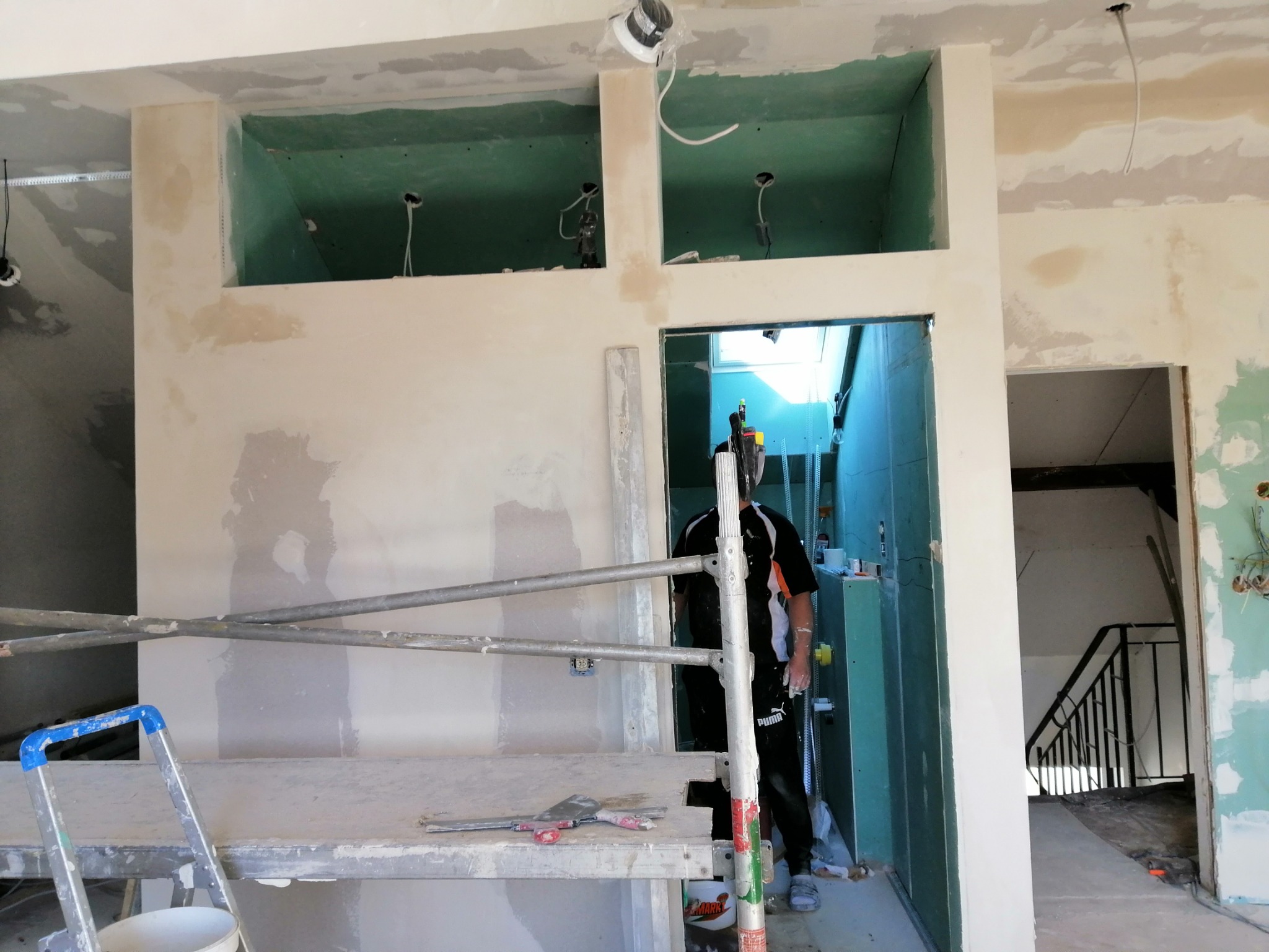 Prace wykończeniowe wnętrza: ściana z otworami pod oświetlenie po szpachlowaniu, widoczne rusztowanie i narzędzia, pracownik w tle.