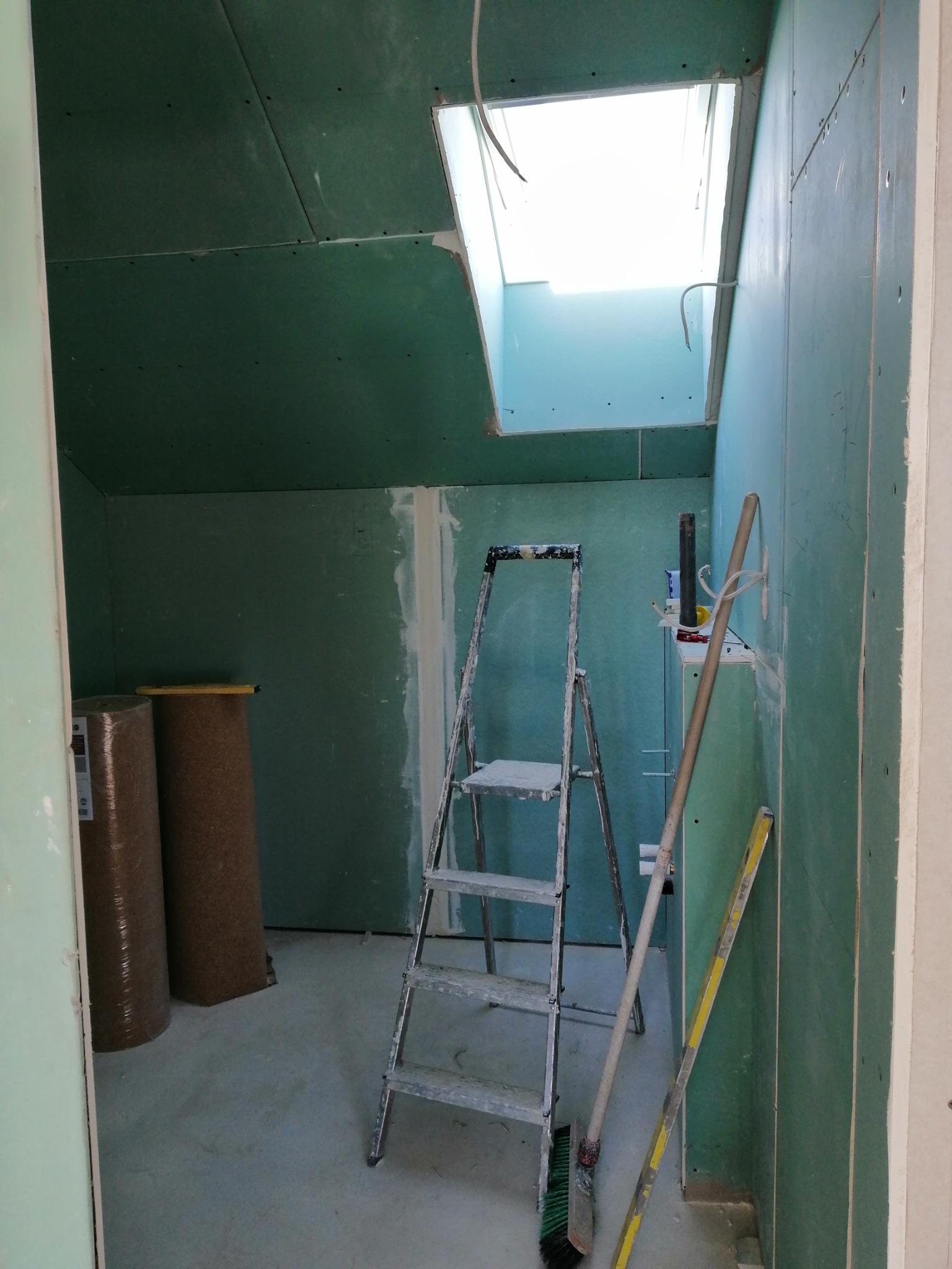 Wnętrze w trakcie wykańczania z surowymi ścianami z płyt gipsowo-kartonowych, widoczne dwa cylindryczne opakowania materiału izolacyjnego, aluminiowa drabina, poziomica i otwór okienny w dachu.