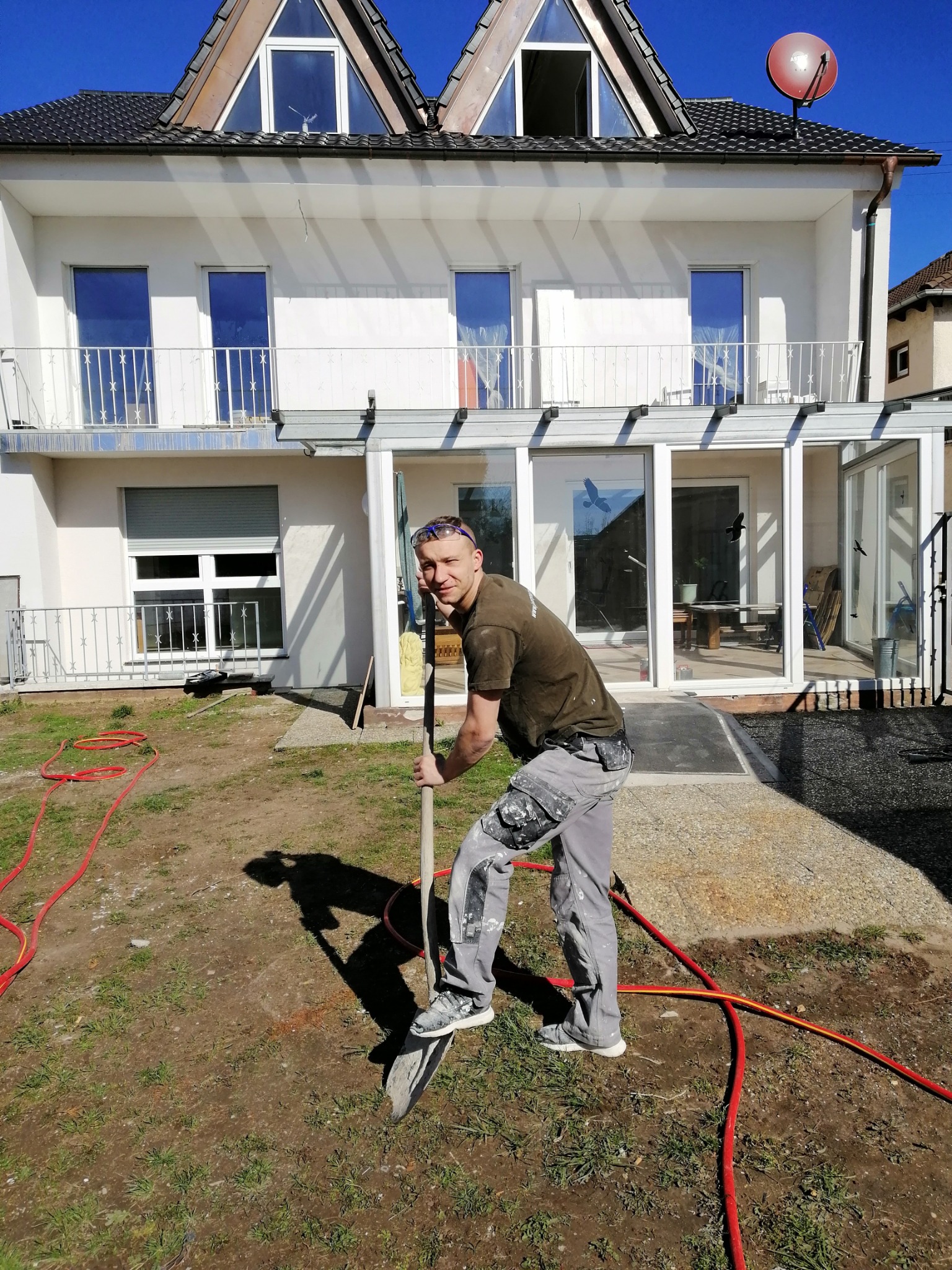 Mężczyzna z łopatą na tle ocieplonego domu z werandą, przygotowujący teren pod prace ziemne, widoczne ślady farby na spodniach i węże ogrodowe na trawniku.