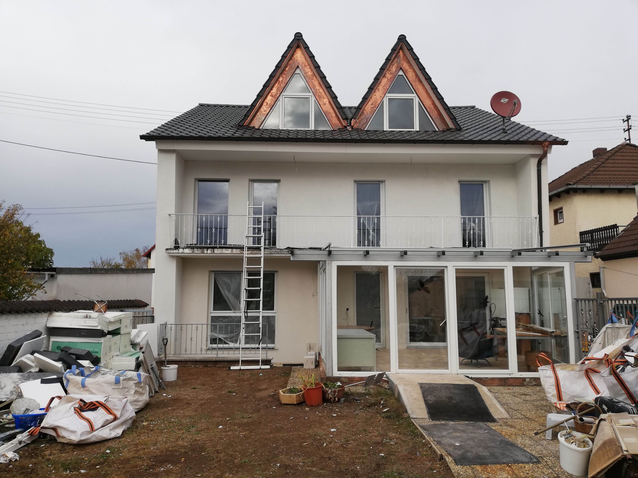 Dwupiętrowy dom z białymi ścianami i dwoma trójkątnymi lukarnami z miedzianym obramowaniem, widoczna drabina oparta o balkon, sterta materiałów budowlanych i worków z odpadami na podwórku, co...
