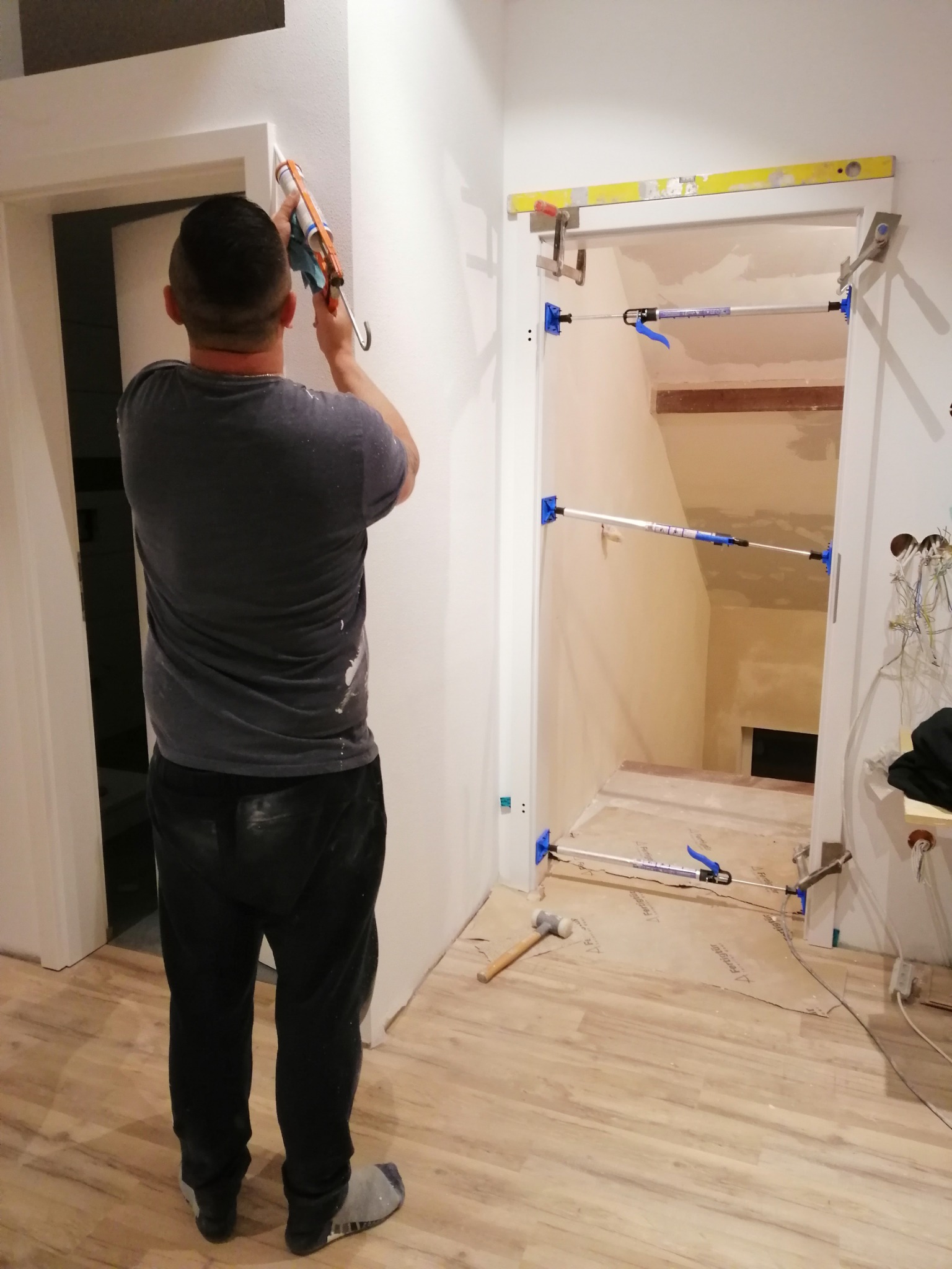 Mężczyzna w szarej koszulce i ciemnych spodniach uszczelnia silikonem ościeżnicę drzwiową, widoczne rozpórki teleskopowe utrzymujące ramę oraz poziomica na górnej belce, podłoga zabezpieczona...