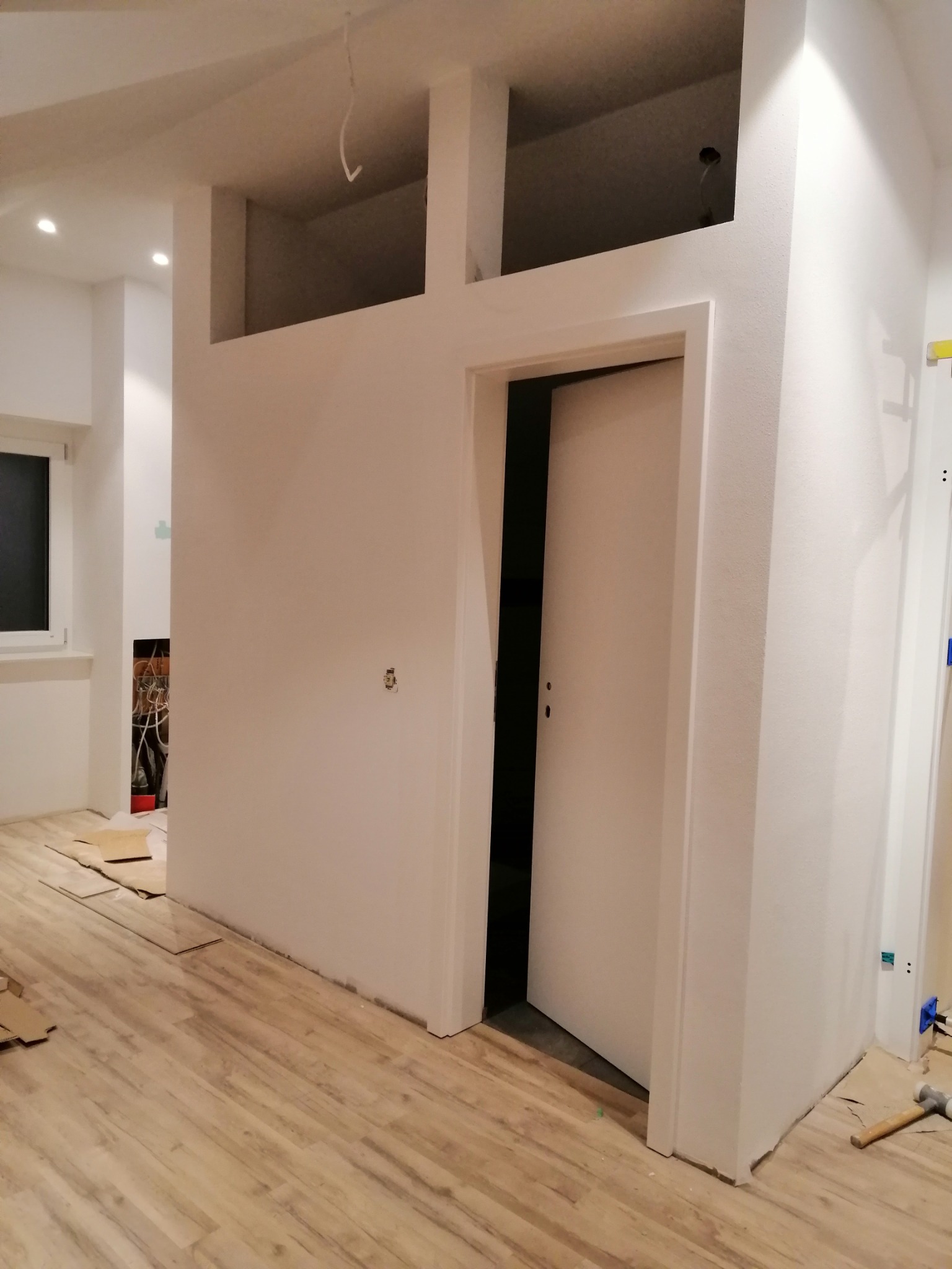 Wnętrze w trakcie remontu: świeżo ułożona podłoga z paneli, pomalowane ściany z wnękami, częściowo otwarte białe drzwi, widoczne narzędzia i materiały budowlane.