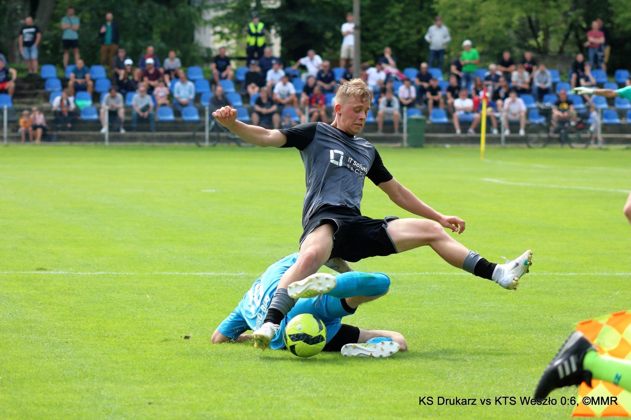 Piłkarz w szarej koszulce i czarnych spodenkach w trakcie skoku nad bramkarzem leżącym na murawie, w tle widownia na trybunach.