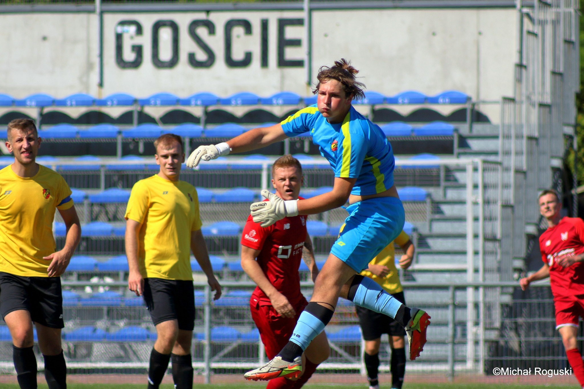 Bramkarz w niebiesko-żółtym stroju w wyskoku podczas interwencji na tle trybun i innych graczy.