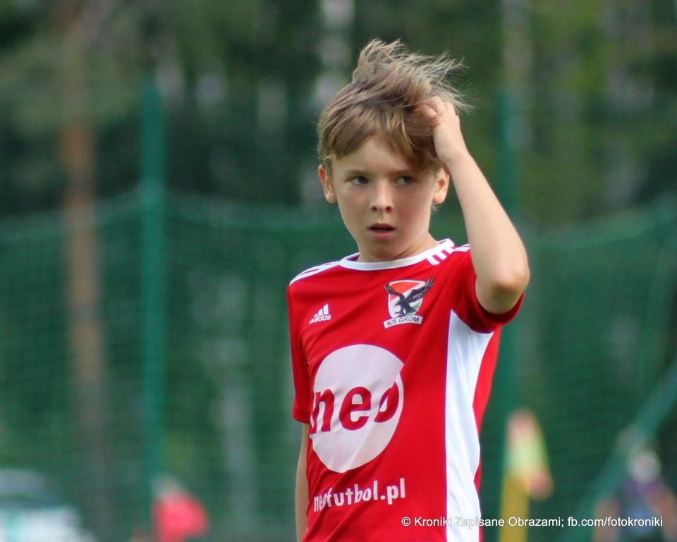 Młody piłkarz w czerwonej koszulce z logo drużyny KS Grom, poprawia włosy, zamyślony wyraz twarzy, na tle zielonej trawy.