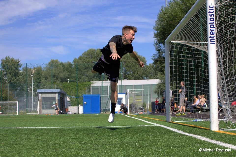 Mężczyzna z tatuażami w czarnym stroju skacze przed bramką na boisku w Warszawie, uchwycony w dynamicznym ujęciu podczas gry w piłkę nożną.