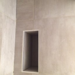 Konrad Stępień - Wnęka prysznicowa wyłożona jasnymi, matowymi płytkami ceramicznymi, widoczna z boku, z delikatnym oświetleniem wewnątrz.