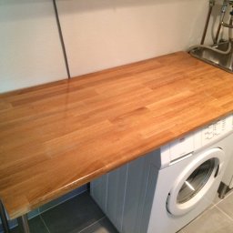 Konrad Stępień - Blat z drewna klejonego nad pralką w łazience, z widocznym zlewem i baterią, oraz szarą podłogą z płytek.