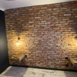 Ściana z czerwonej cegły z zamontowanymi półkami nocnymi i lampami w stylu industrialnym, na jednej półce leży pilot i kabel USB.
