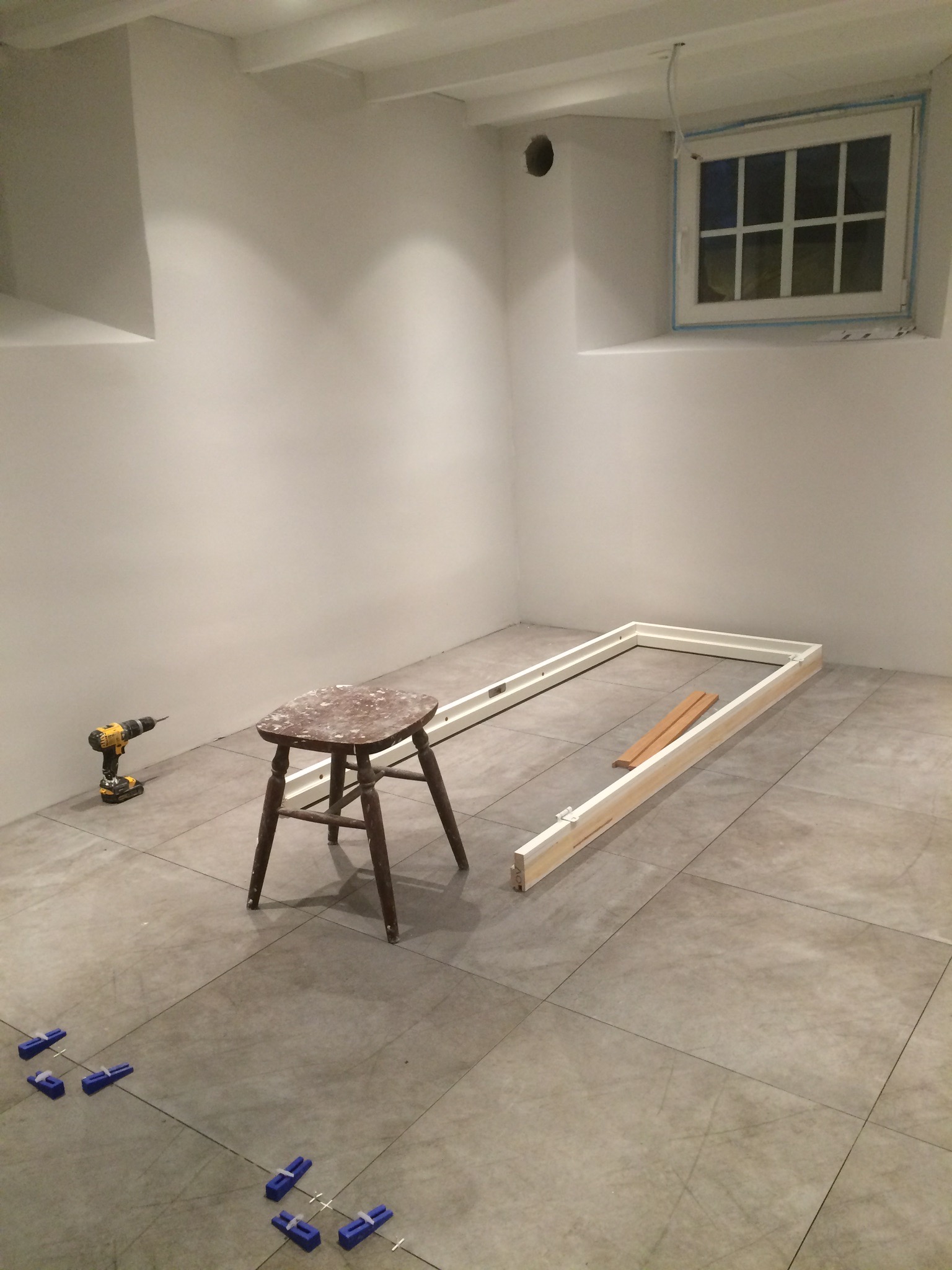 Przygotowania do montażu ościeżnicy w surowym pomieszczeniu z szarymi płytkami podłogowymi, widoczna wiertarka akumulatorowa, drewniany stołek i niebieskie kliny dystansowe do płytek.