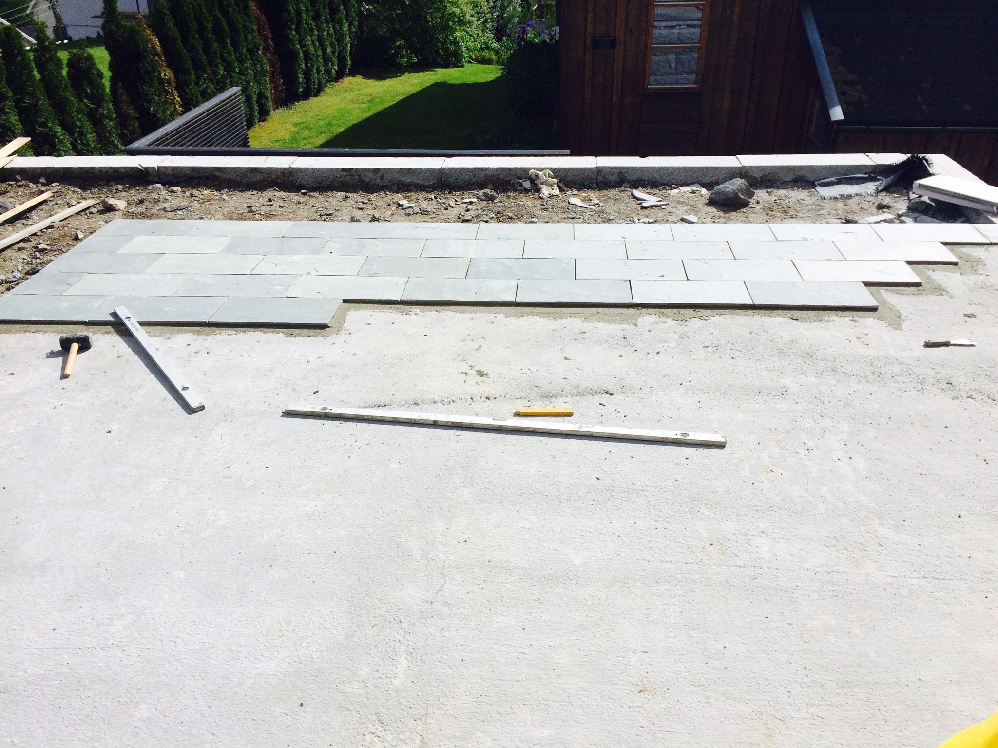 Układanie szarych płytek na tarasie zewnętrznym, widoczny młotek, poziomica i fragmenty ogrodu w tle.