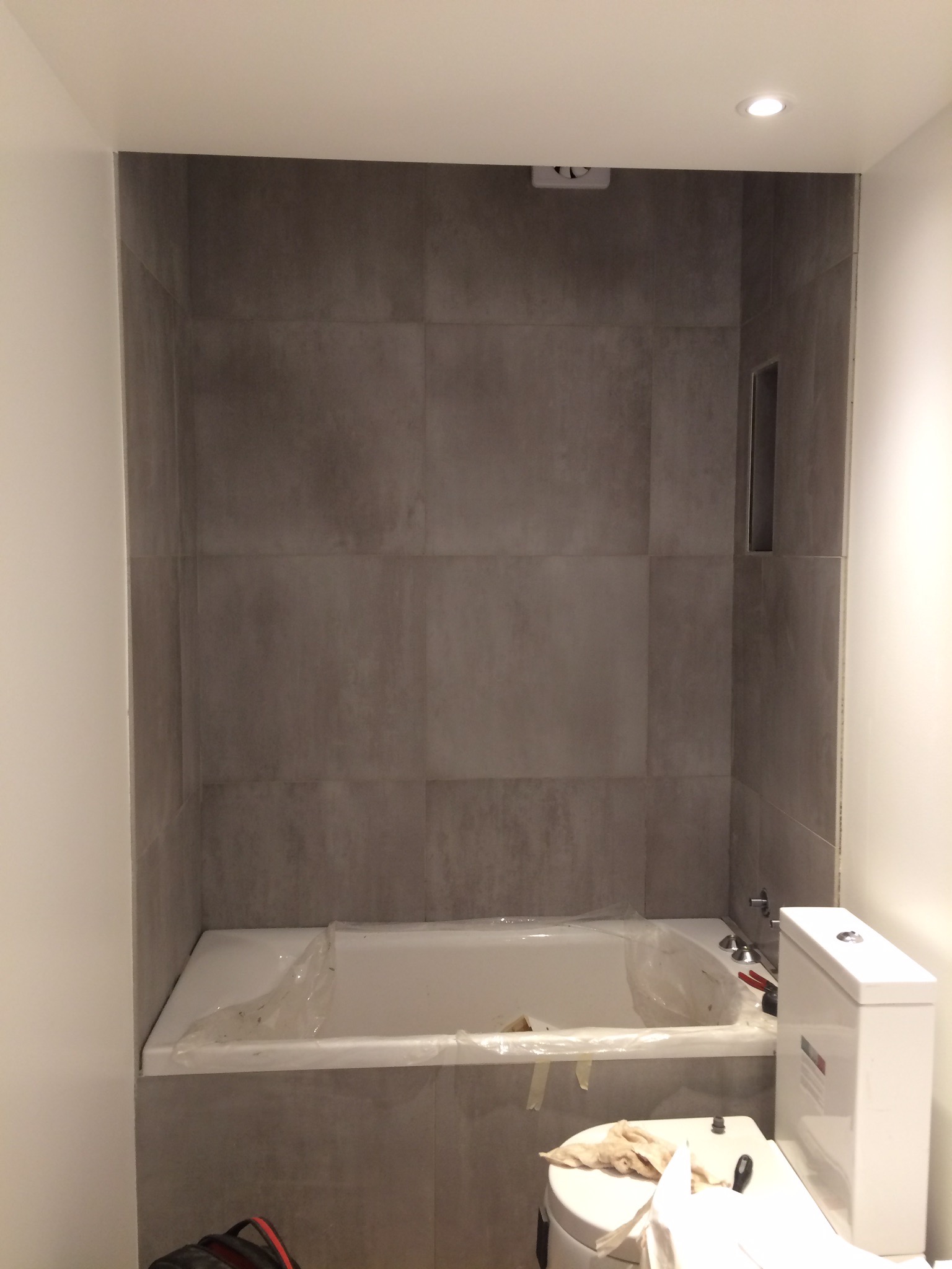 Szara glazura ścienna w trakcie układania nad wanną, częściowo zabezpieczona folią, widoczna instalacja toalety w tle, oświetlenie sufitowe.
