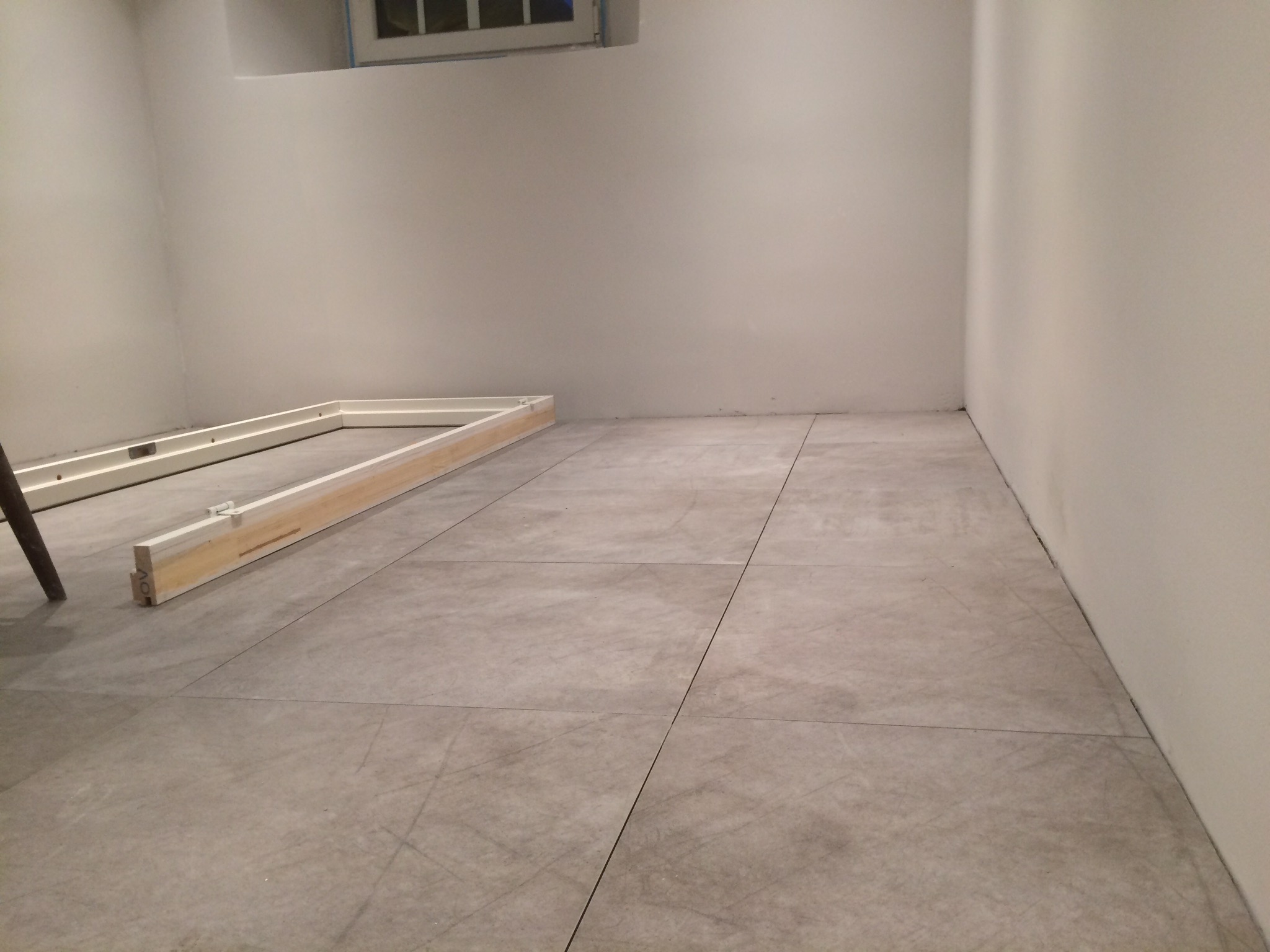 Szare płytki podłogowe ułożone w kwadraty, z widocznymi fugami i drewnianą konstrukcją ramy drzwi w rogu pomieszczenia, ściany pomalowane na biało.