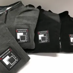 Rząd polarowych bluz z zamkiem błyskawicznym w odcieniach szarości i czerni, z naszytym logo 'Ekonomiczne Instalacje' w czarno-białej kolorystyce.