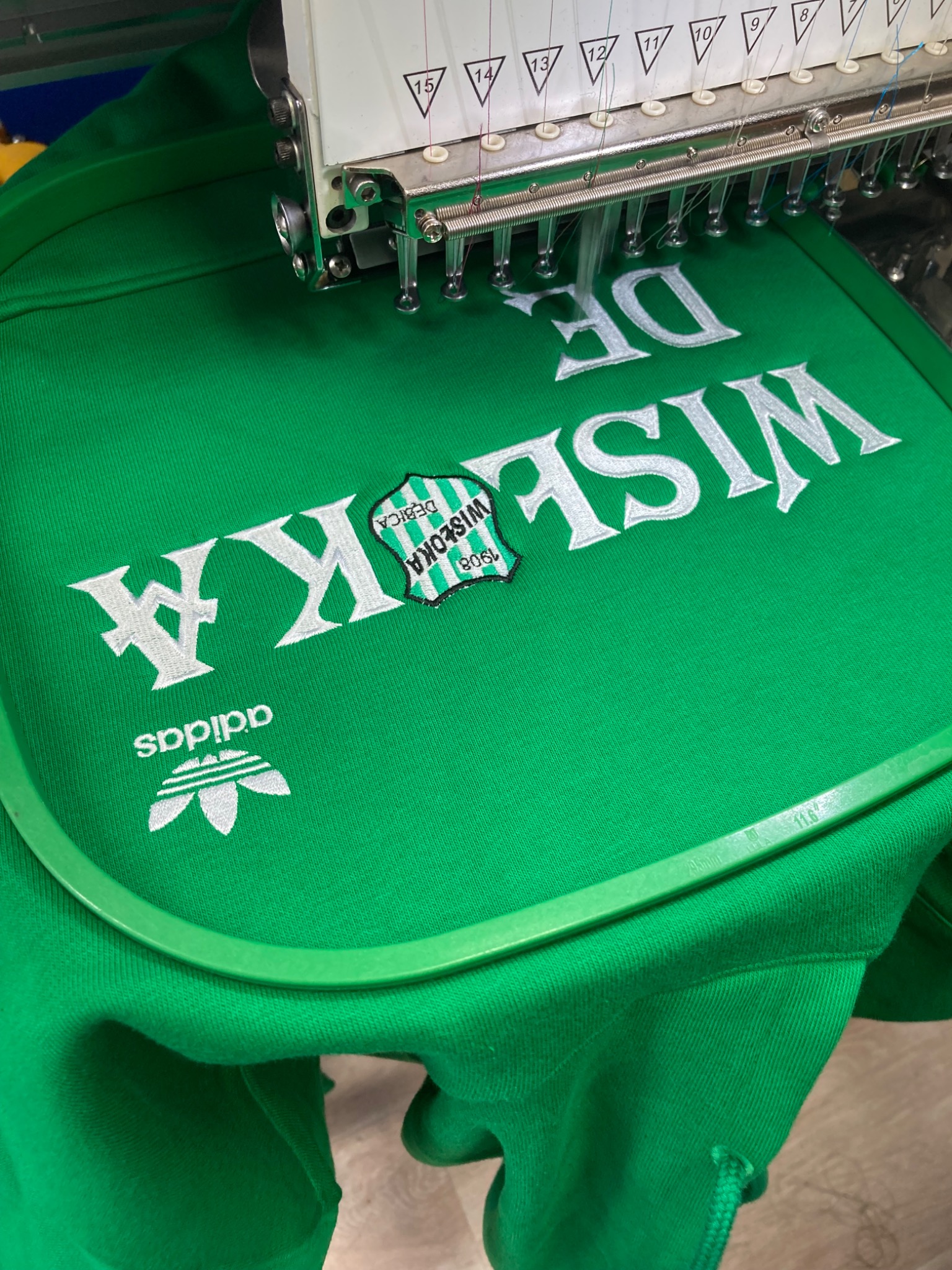 Proces haftowania napisu 'Wisłoka Dębica' oraz logo Adidas na zielonej bluzie, widoczna głowica maszyny hafciarskiej z nawleczonymi nićmi.