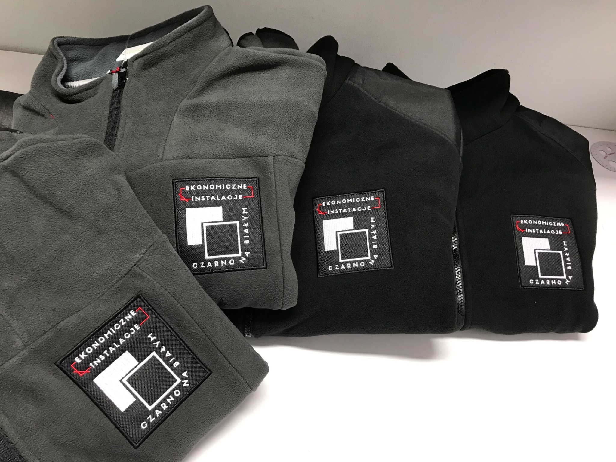 Rząd polarowych bluz z zamkiem błyskawicznym w odcieniach szarości i czerni, z naszytym logo 'Ekonomiczne Instalacje' w czarno-białej kolorystyce.