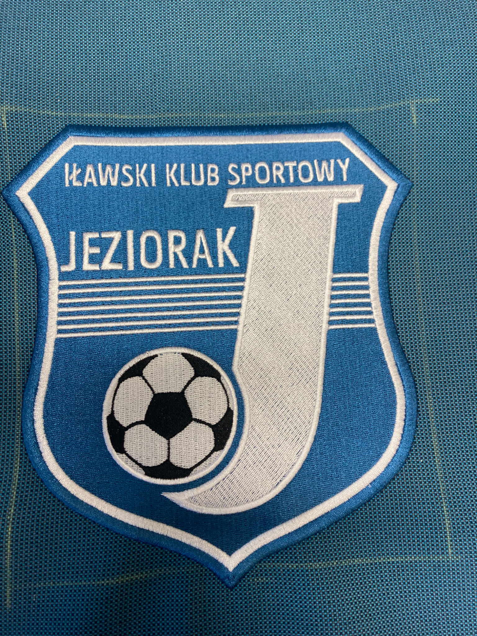 Niebiesko-biała naszywka z wyhaftowanym herbem Iławskiego Klubu Sportowego Jeziorak, przedstawiająca piłkę nożną i literę J na tle tkaniny.