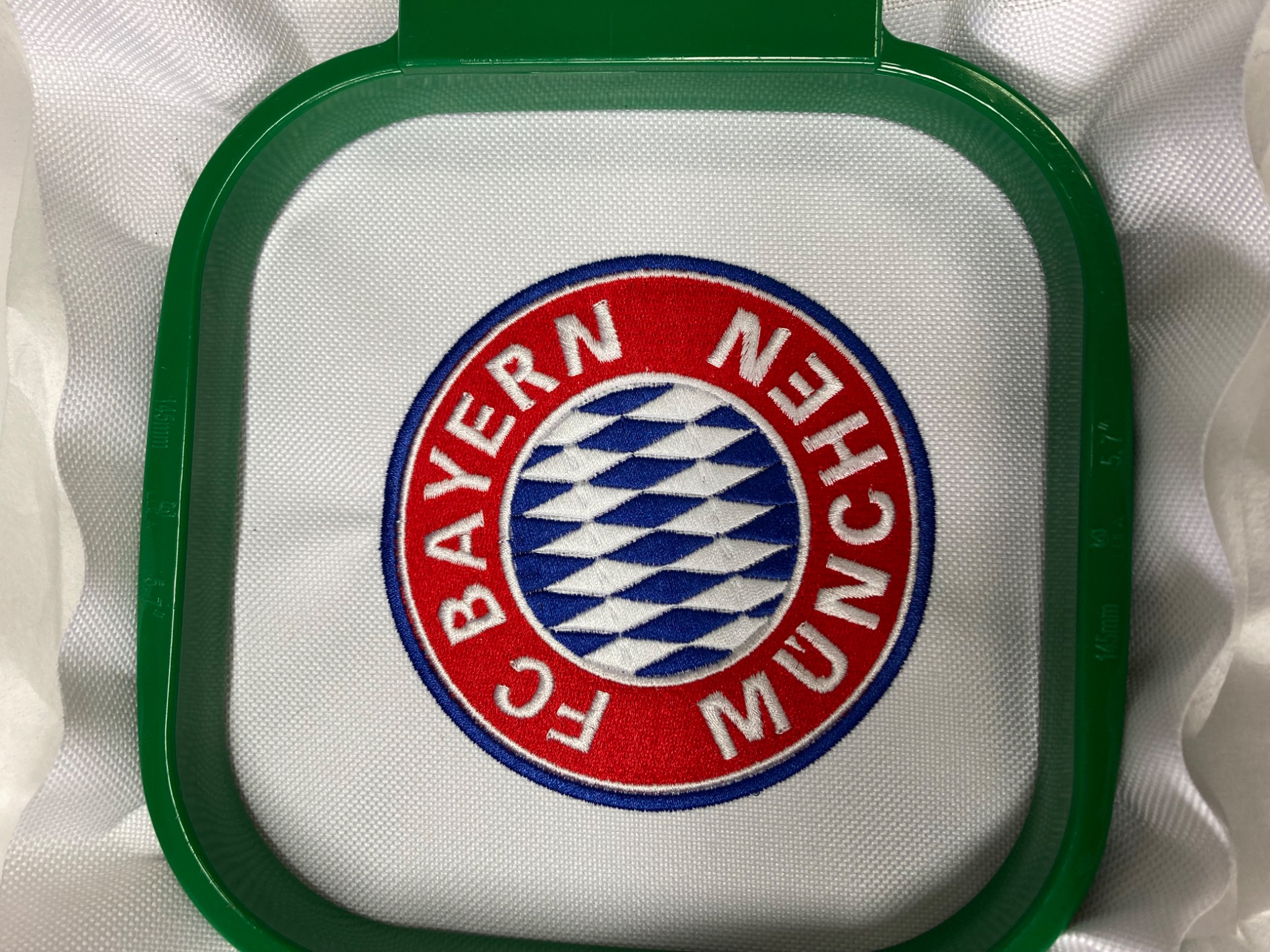 Emblemat FC Bayern Monachium wyhaftowany na białym materiale, umieszczony w zielonej ramce do haftowania.