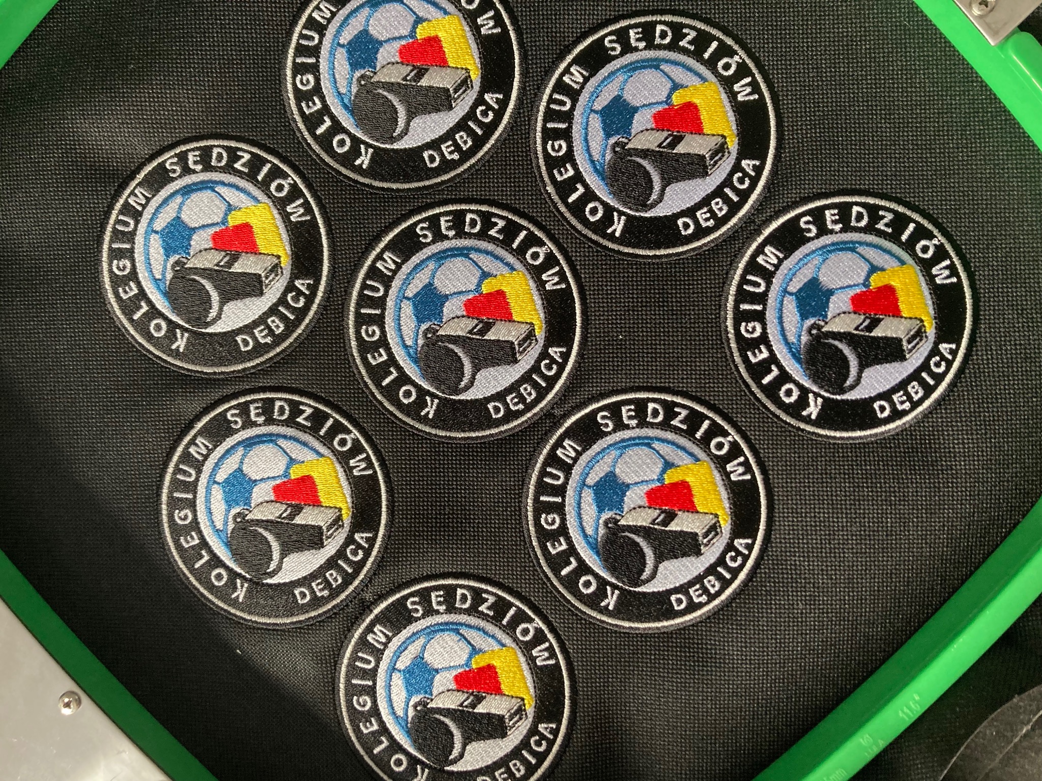 Seria okrągłych naszywek z haftowanym logo 'Kolegium Sędziów Dębica' przedstawiającym piłkę nożną, gwizdek sędziowski i flagi w kolorach żółtym, czerwonym i niebieskim, ułożonych na czarnym...