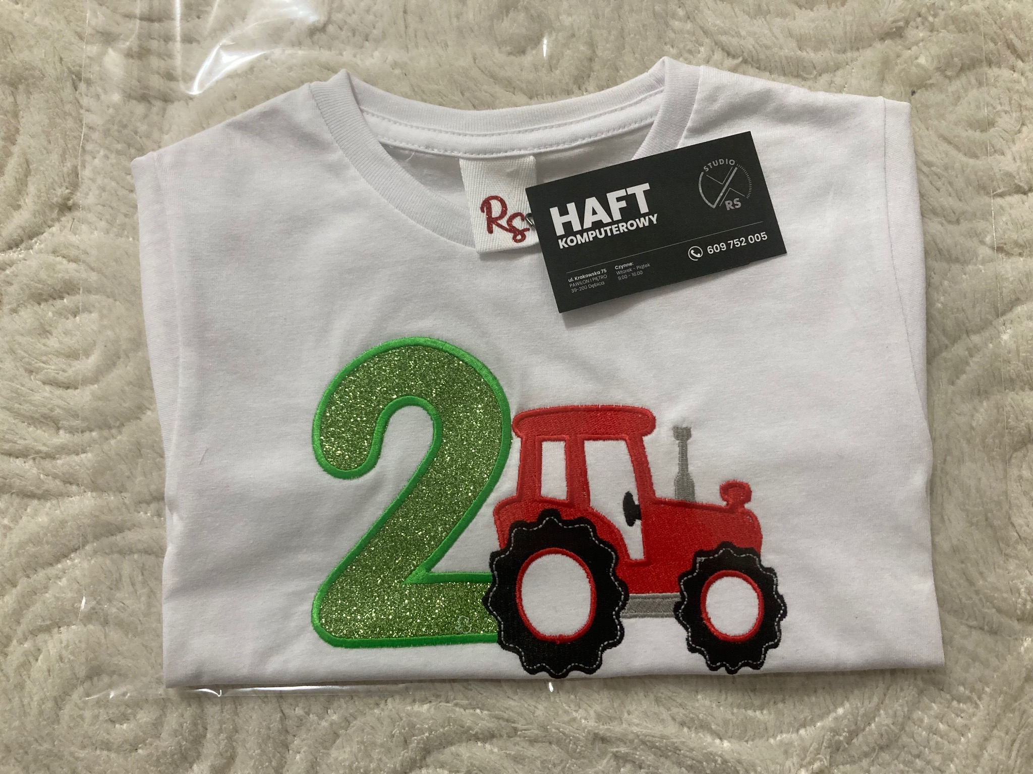 Biała koszulka dziecięca z haftowanym, brokatowym numerem '2' i czerwonym traktorem, leżąca na puszystym tle; widoczna metka i wizytówka firmy oferującej usługi komputerowe.