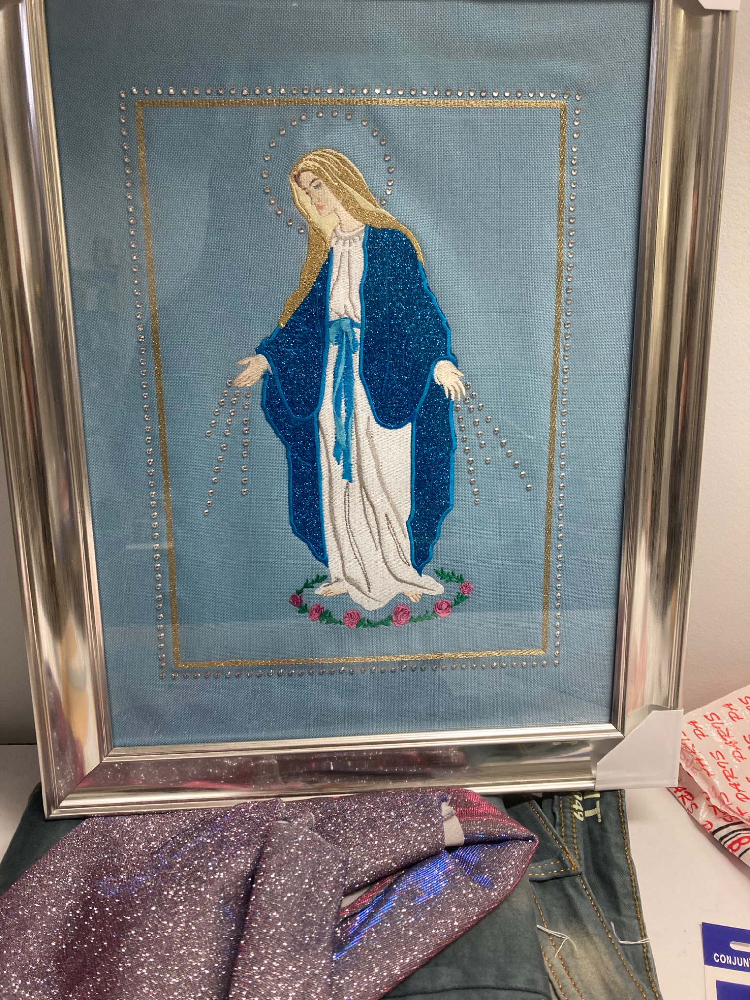 Oprawiony w srebrną ramę, haftowany obraz Matki Boskiej na błękitnym tle, ozdobiony koralikami i złotą nicią, leżący na stole obok błyszczącej tkaniny i dżinsów.