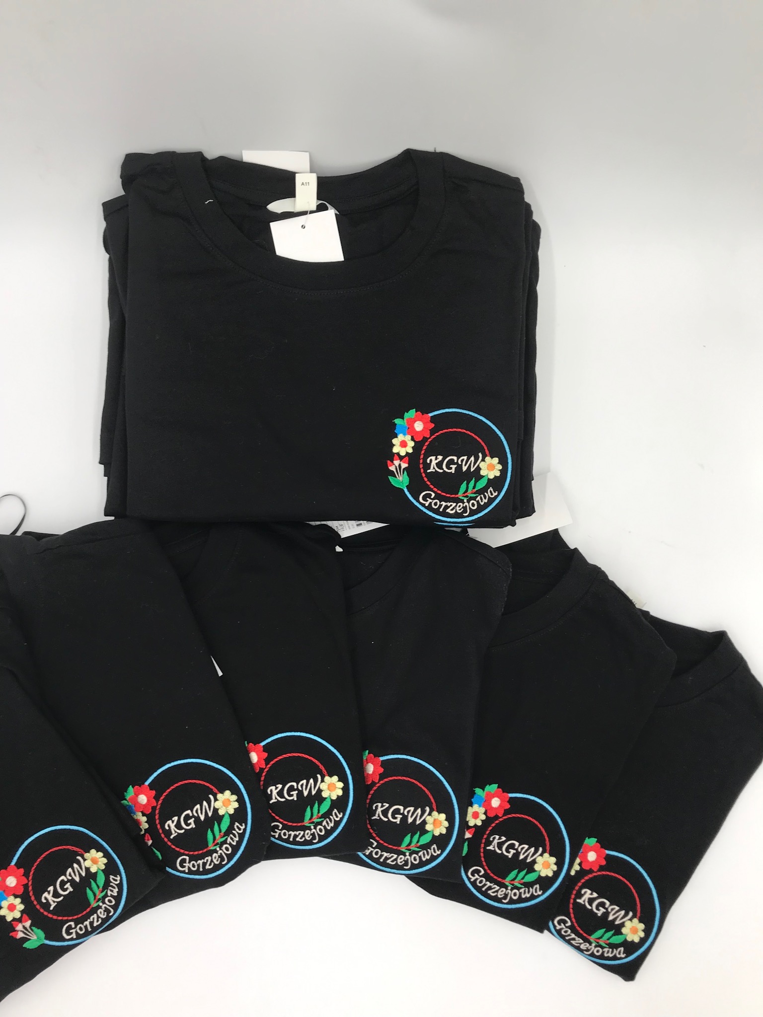 Stos czarnych koszulek z haftowanym logo KGW Gorzejowa otoczonym kwiatami i niebieskim okręgiem, ułożonych na białym tle. Na górnej koszulce widoczna metka.