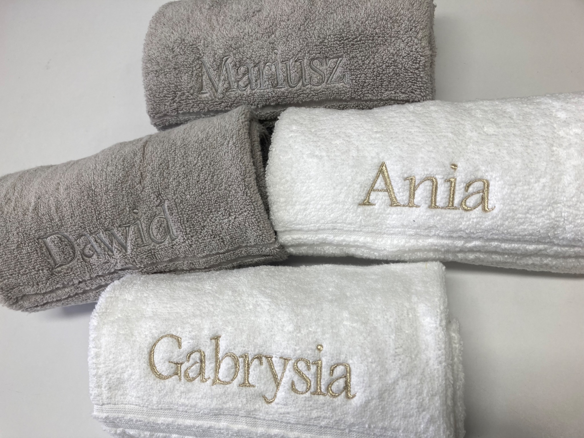 Stos czterech ręczników: dwa szare z imionami 'Mariusz' i 'Dawid' wyhaftowanymi szarą nicią oraz dwa białe z imionami 'Ania' i 'Gabrysia' wyhaftowanymi złotą nicią, ułożone na białym tle.