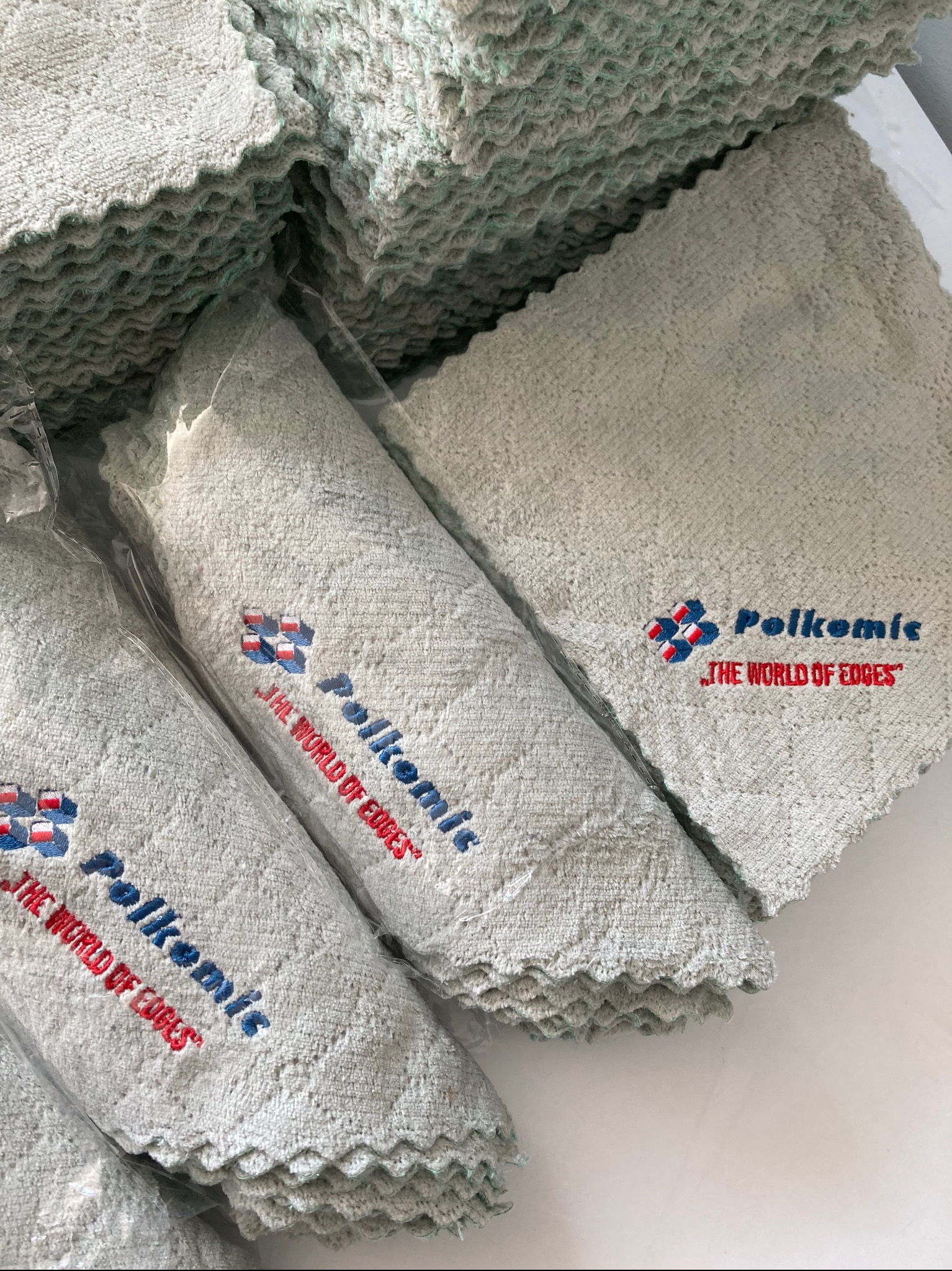 Stos szarych ręczników z wyhaftowanym logo firmy 'Polkemis', część z nich zapakowana w folię.