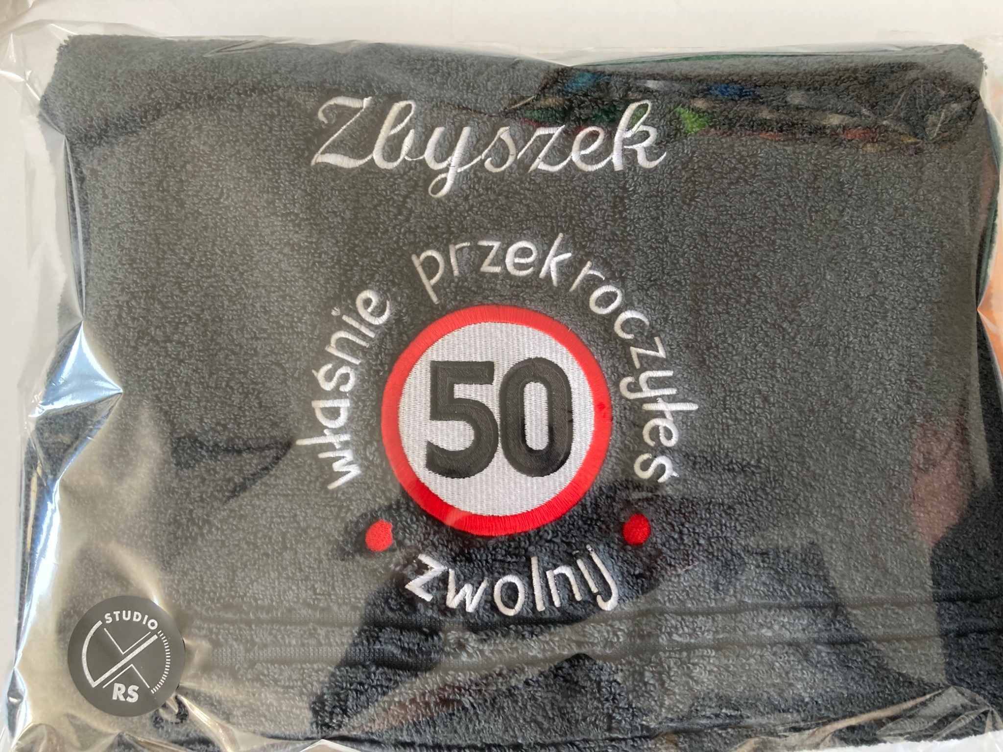 Szary ręcznik zapakowany w folię z białym haftem: imię 'Zbyszek' oraz grafika koła z liczbą 50 i napisem 'właśnie przekroczyłeś, zwolnij'.