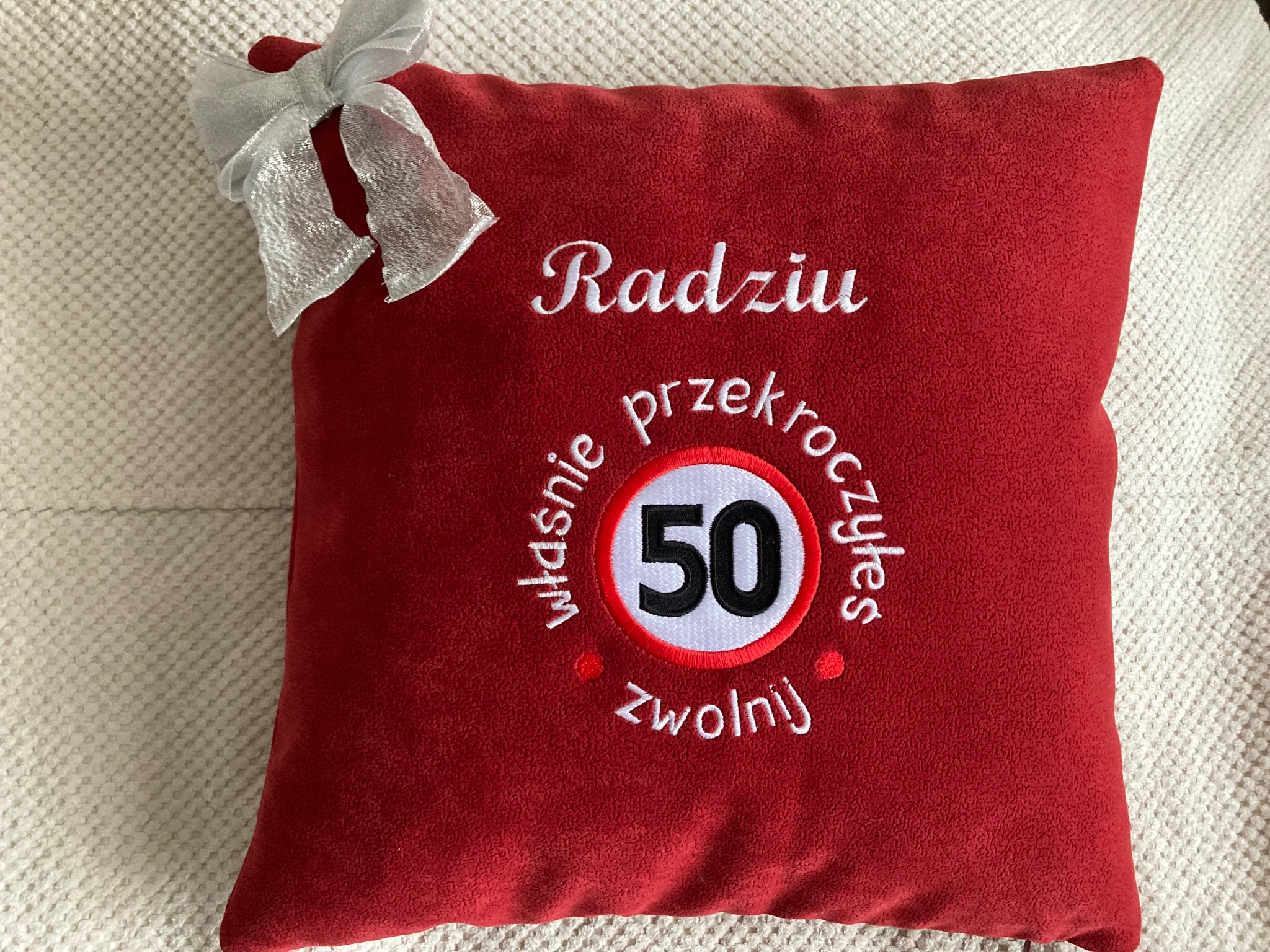 Czerwona poduszka z białym haftem 'Radziu właśnie przekroczyłeś 50 zwolnij' i symbolem ograniczenia prędkości, ozdobiona srebrną kokardą w lewym górnym rogu.