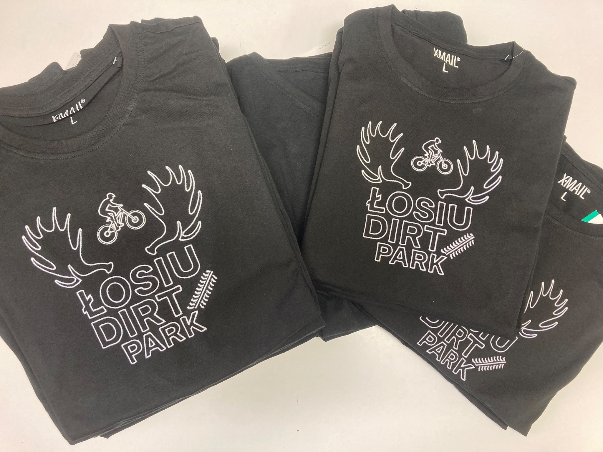 Stos czarnych koszulek z białym nadrukiem 'Łosiu Dirt Park' i motywem rowerzysty w porożu. Widoczne metki 'XMAIL' i 'KAAALL'.