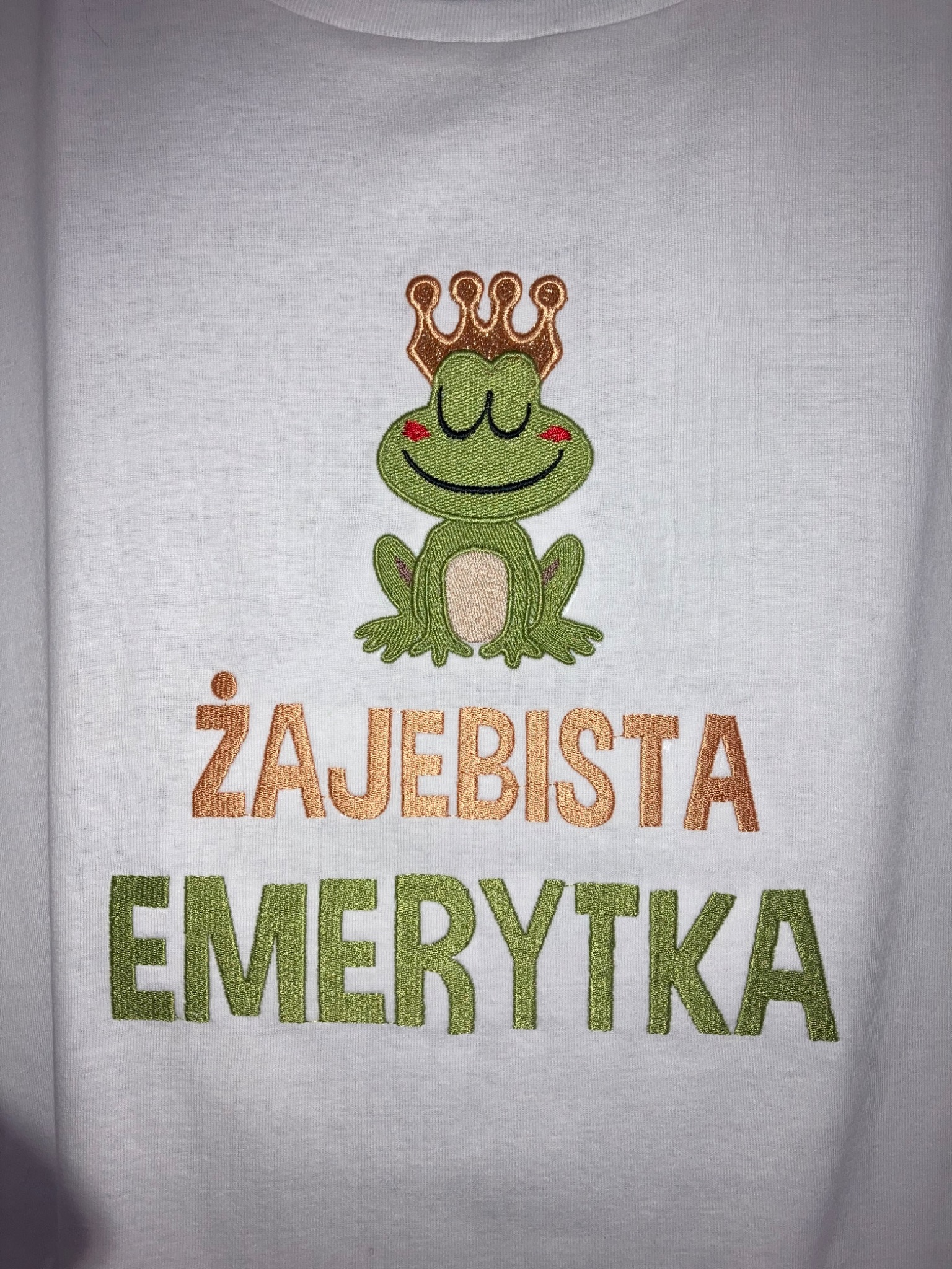 Biała koszulka z haftowanym motywem żaby w koronie i napisem 'ŻAJEBISTA EMERYTKA' w kolorach złotym i zielonym.