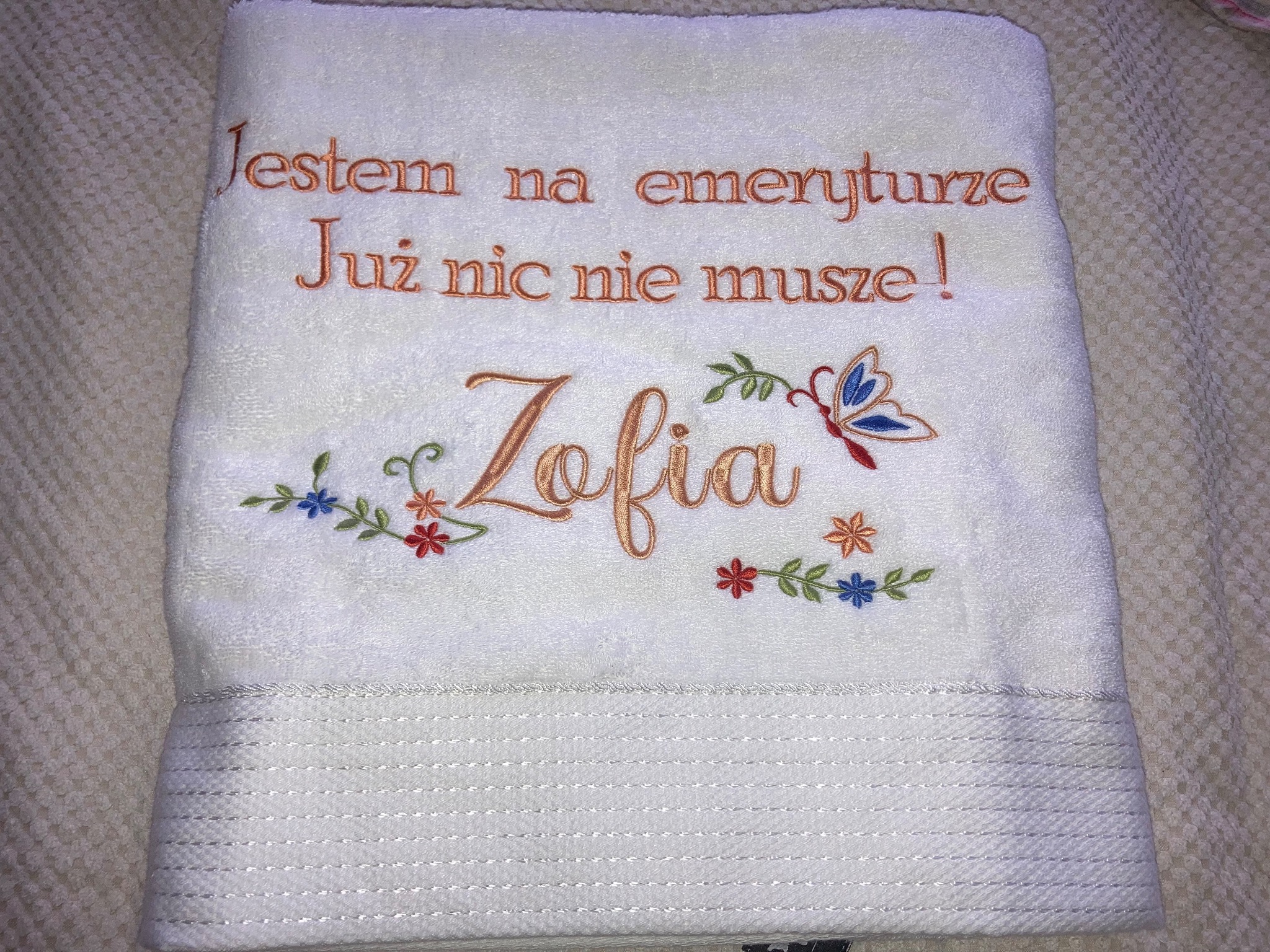 Biały ręcznik z wyhaftowanym napisem 'Jestem na emeryturze. Już nic nie muszę!' i imieniem 'Zofia' ozdobionym motywami kwiatowymi i motylem w kolorach pomarańczowym i niebieskim.