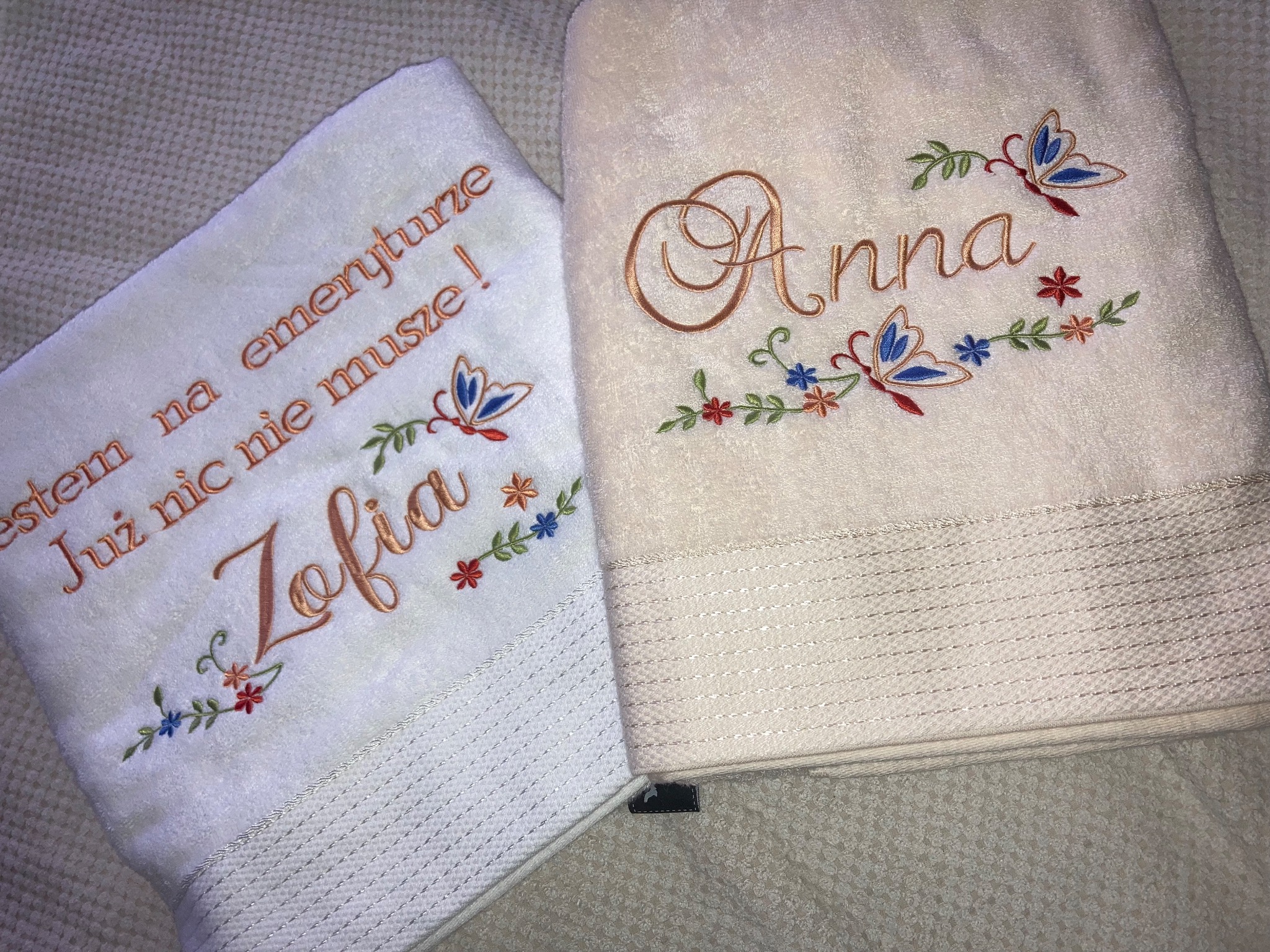 Dwa ręczniki frotte: biały z napisem 'Zofia' i beżowy z napisem 'Anna', oba zdobione motywami kwiatowymi i motylami wykonanymi techniką zdobniczą.