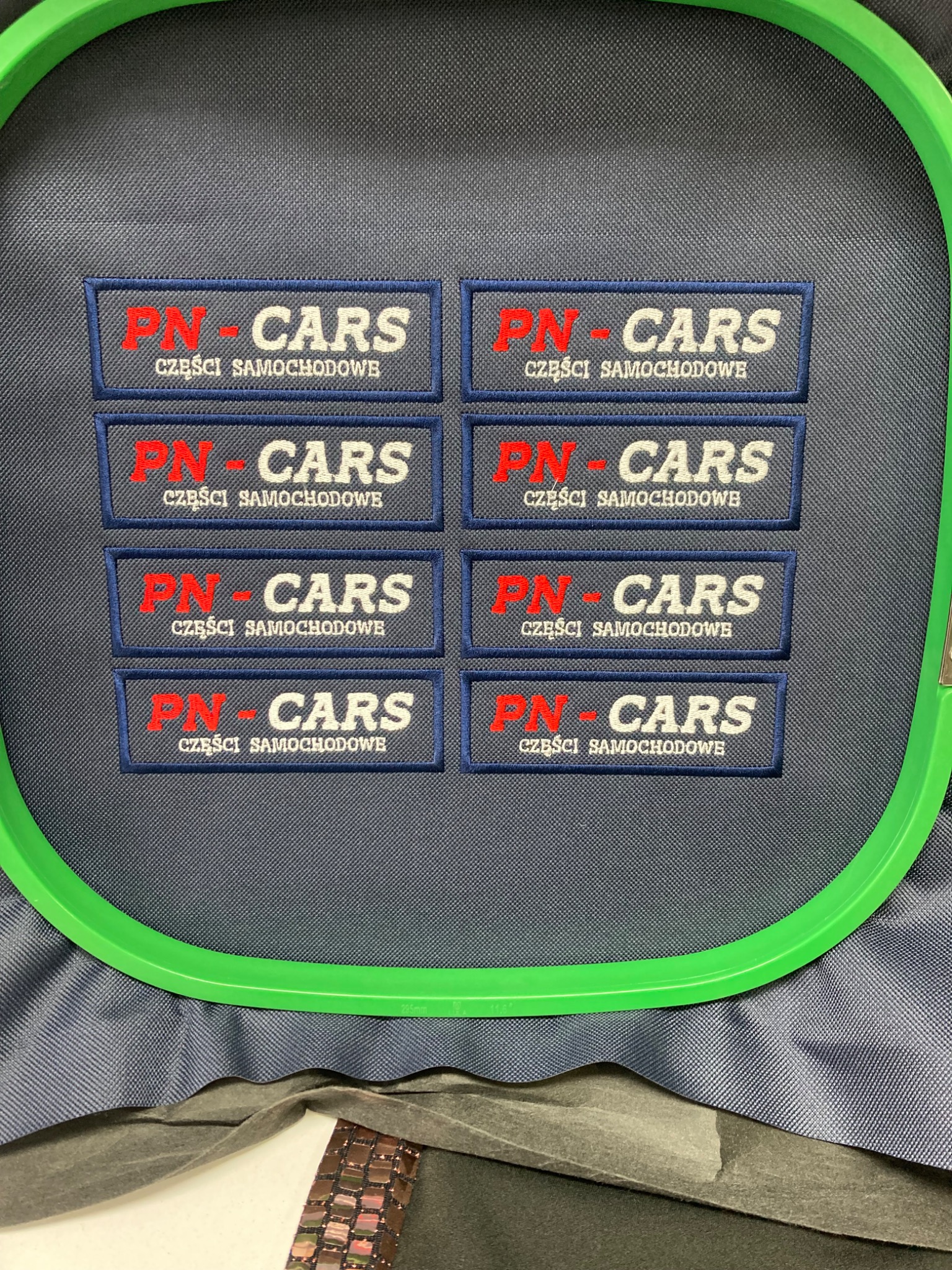 Gotowe naszywki z logo 'PN-CARS' i napisem 'CZĘŚCI SAMOCHODOWE' w ramce,  wykonane techniką  na materiale. Detal pracowni odzieżowej.