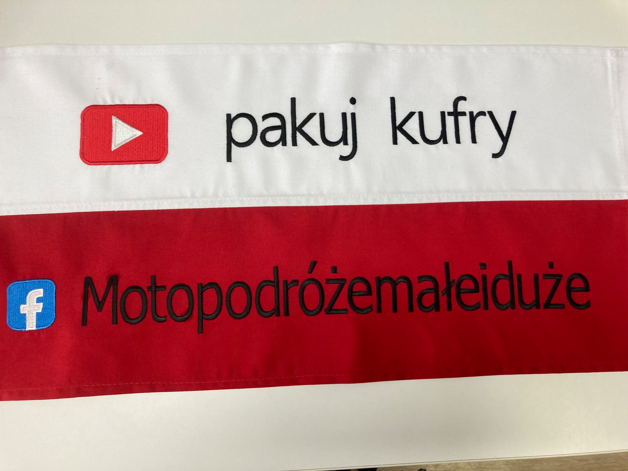 Haftowany baner z logo YouTube i Facebook oraz napisami 'pakuj kufry' i 'Motopodróżemałeiduże' na białym i czerwonym tle, prezentujący precyzję wykonania.