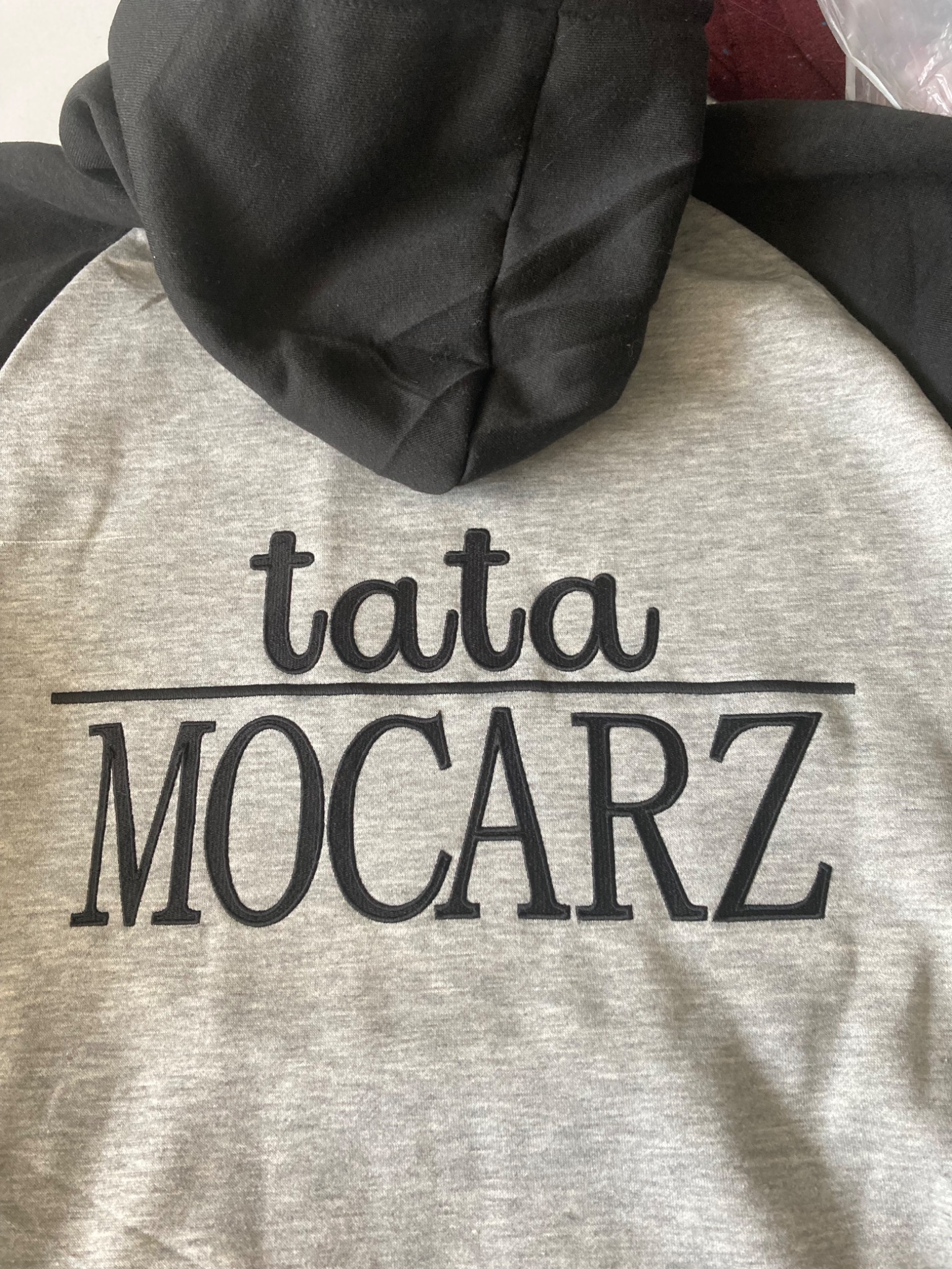 Szara bluza z kapturem i czarnymi rękawami, z wyhaftowanym napisem 'tata MOCARZ' czarną nicią. Widok z tyłu, kaptur częściowo zasłania napis.