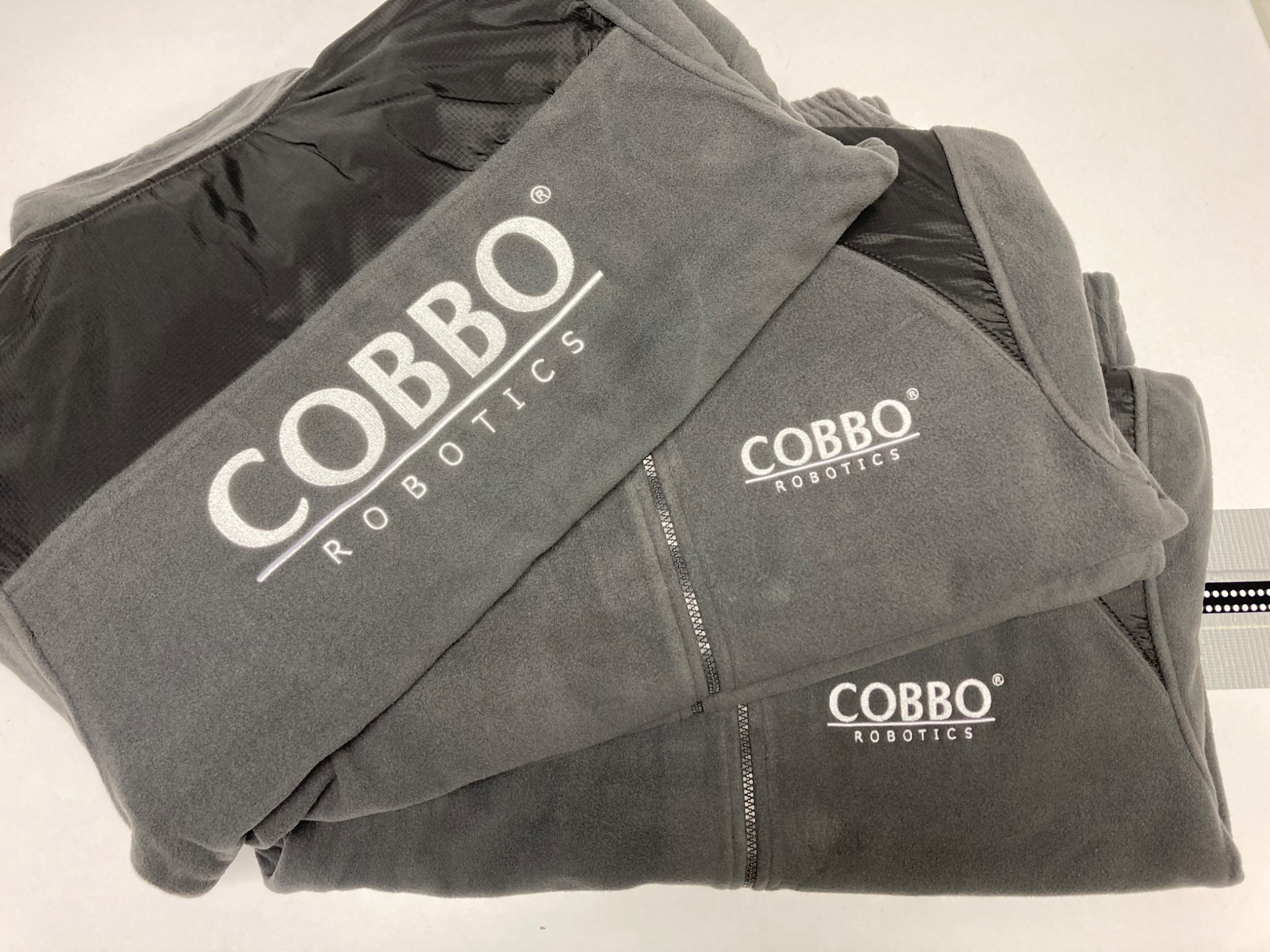 Stos szarych polarów z haftowanym białym logo 'Cobbo Robotics', widoczne detale zamków i materiałów. Precyzyjne wykonanie haftu.