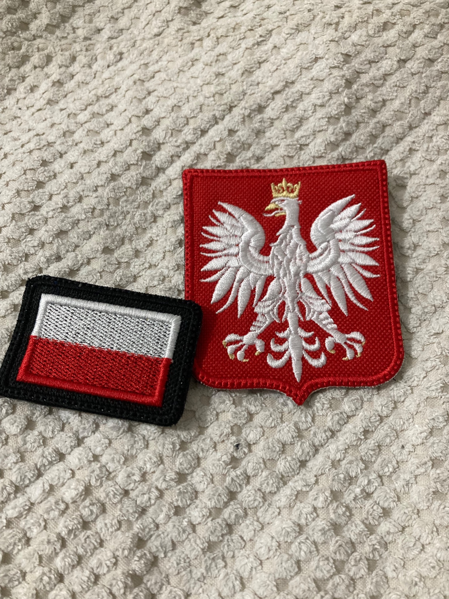 Dwie naszywki: flaga Polski w czarnej obwódce i haftowany orzeł na czerwonym tle, leżą na teksturowanej, kremowej tkaninie. Detal haftu widoczny.