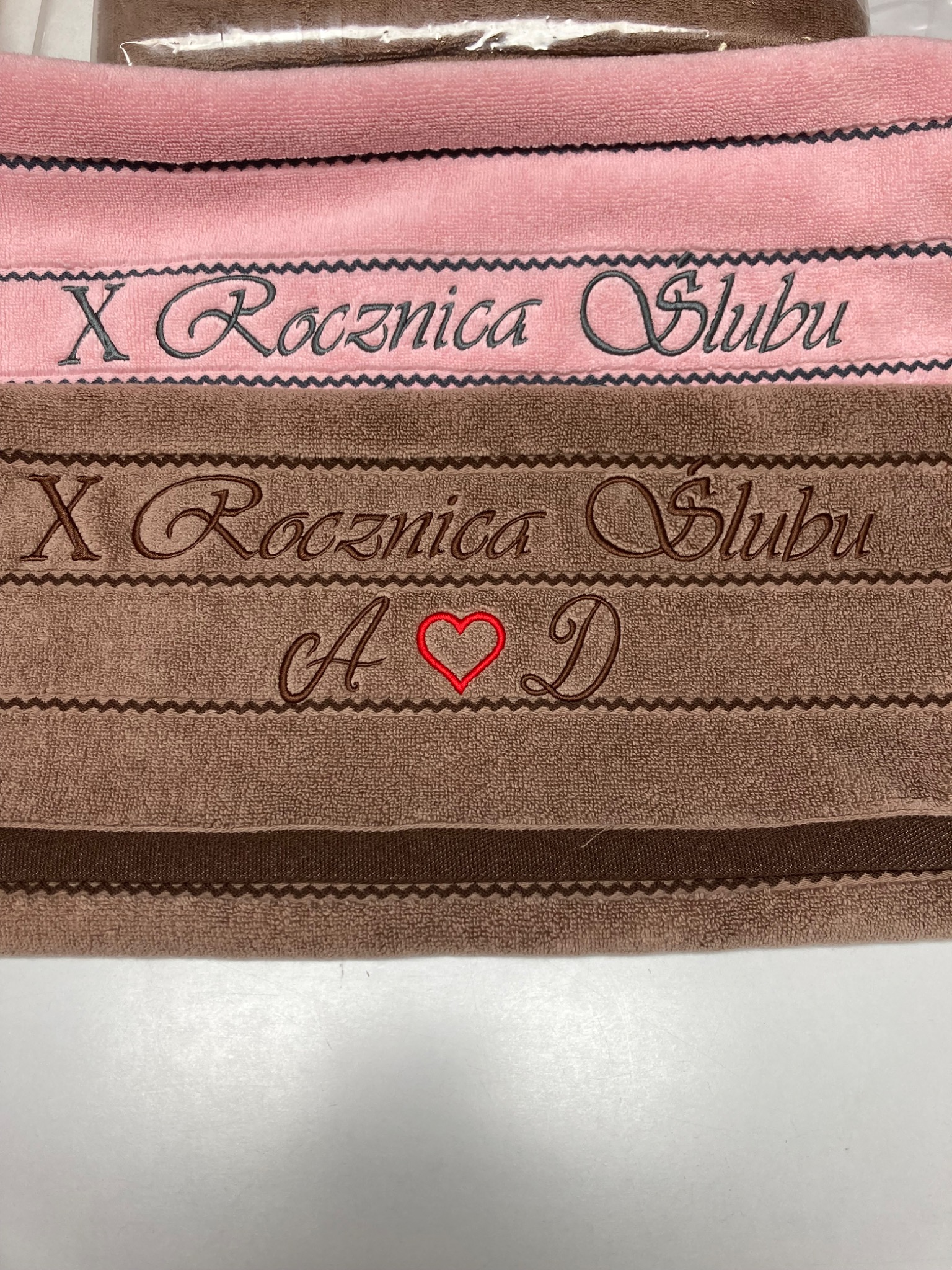 Różowy i brązowy ręcznik z wyhaftowanym napisem 'X Rocznica Ślubu' oraz inicjałami 'A' i 'D' przedzielonymi czerwonym sercem. Detal prezentu.