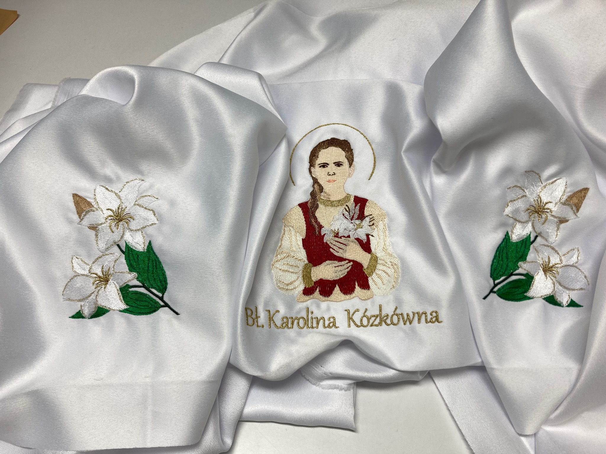Biała tkanina z precyzyjnym, kolorowym zdobieniem przedstawiającym postać Bł. Karoliny Kózkówny i kwiatowe motywy. Złote napisy i delikatne cieniowanie.
